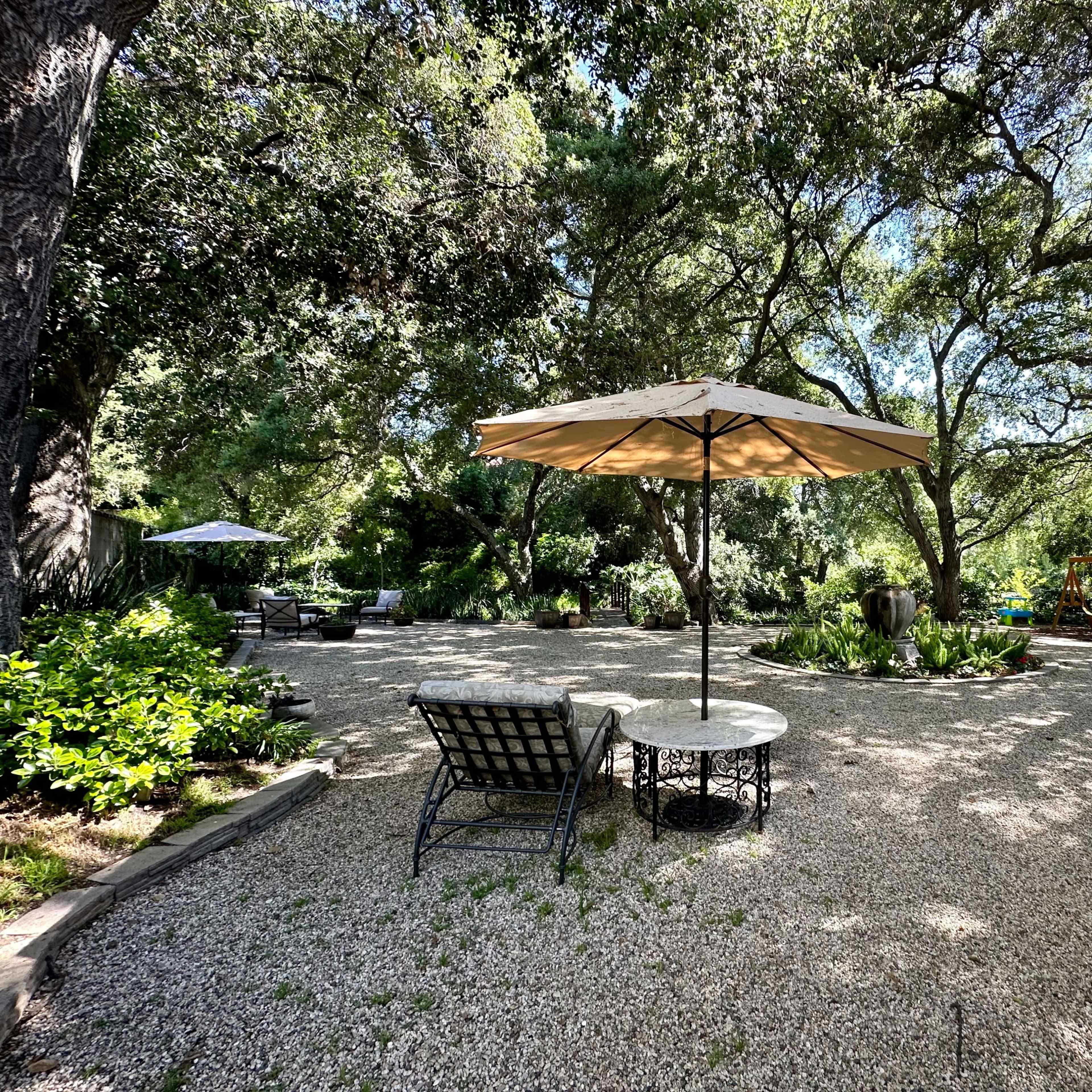 A gravel garden area features a table and chairs under an umbrella, surrounded by lush greenery and trees.