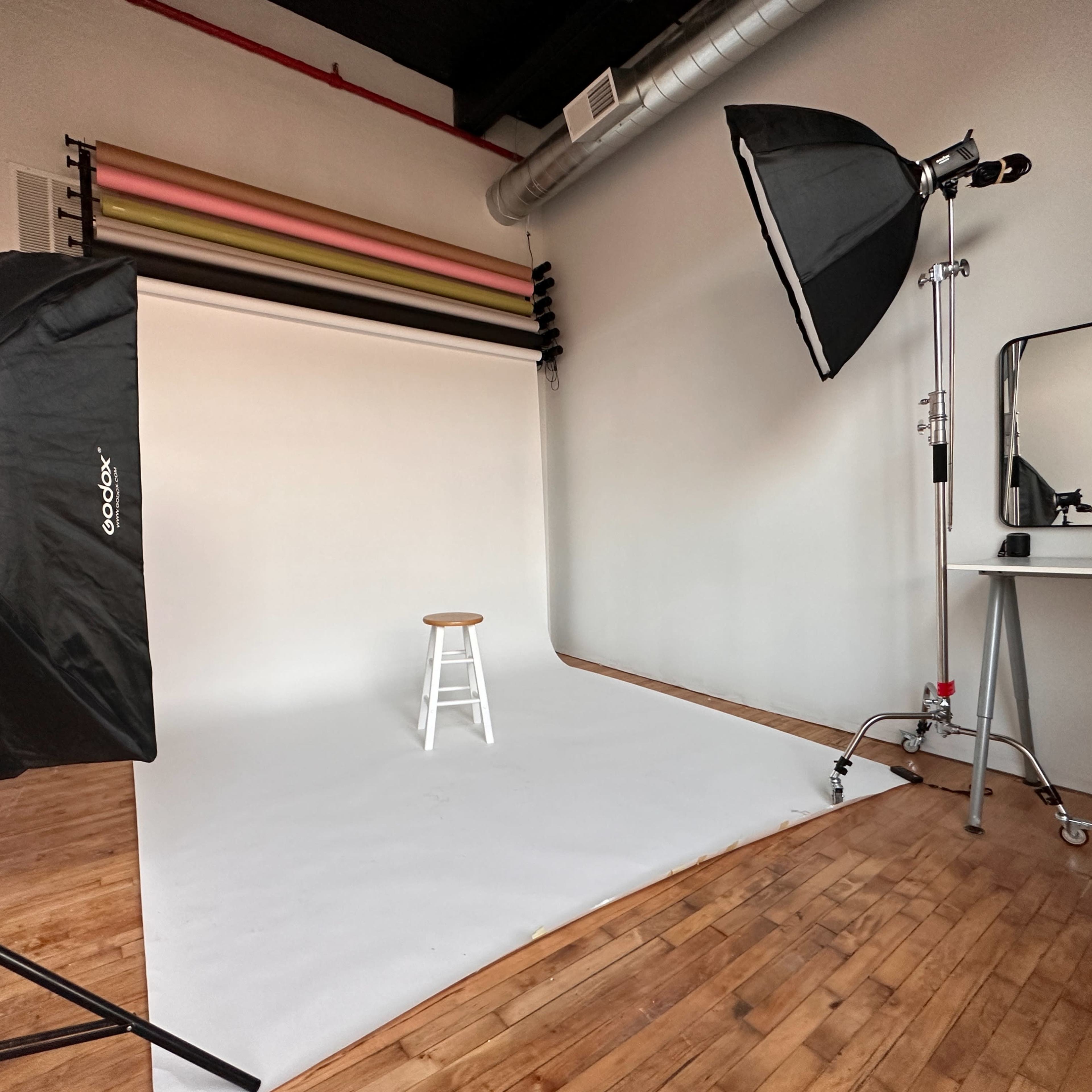 A photo studio setup with a white backdrop, a stool in the center, lighting equipment, and a mirror on the side.
