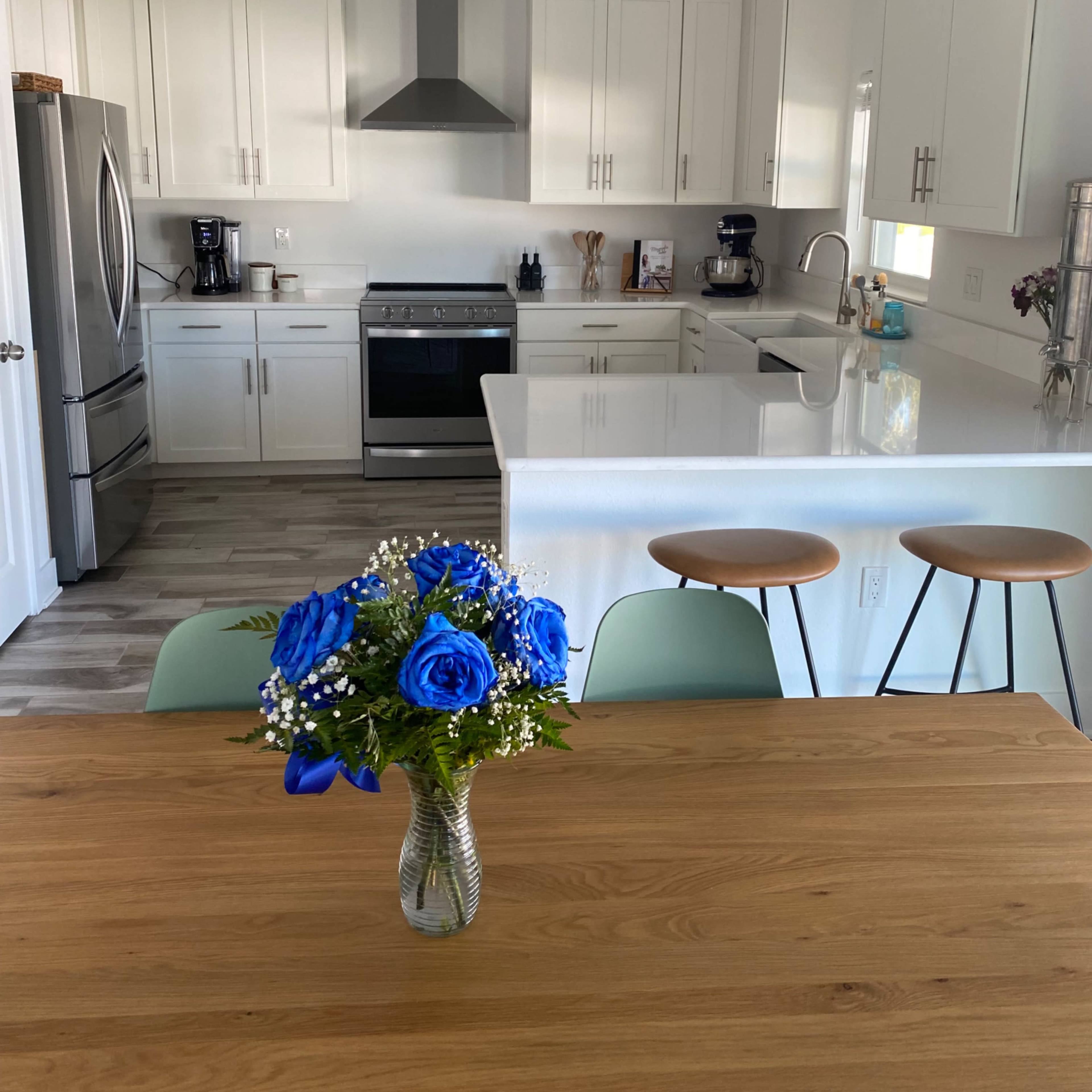A modern kitchen features white cabinetry, stainless steel appliances, a large island with bar stools, and a dining table with a vase of blue roses.