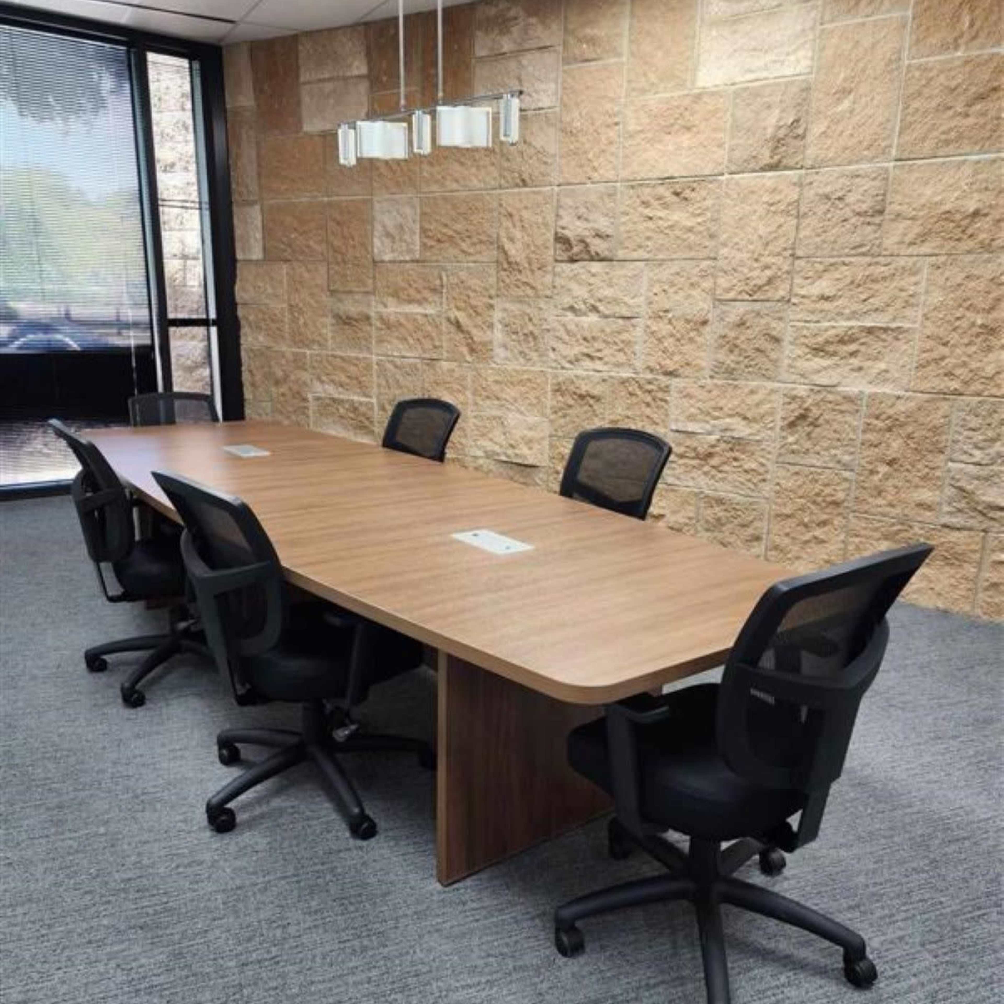 A long wooden conference table with five black office chairs is positioned in a meeting room with a stone wall and large windows.