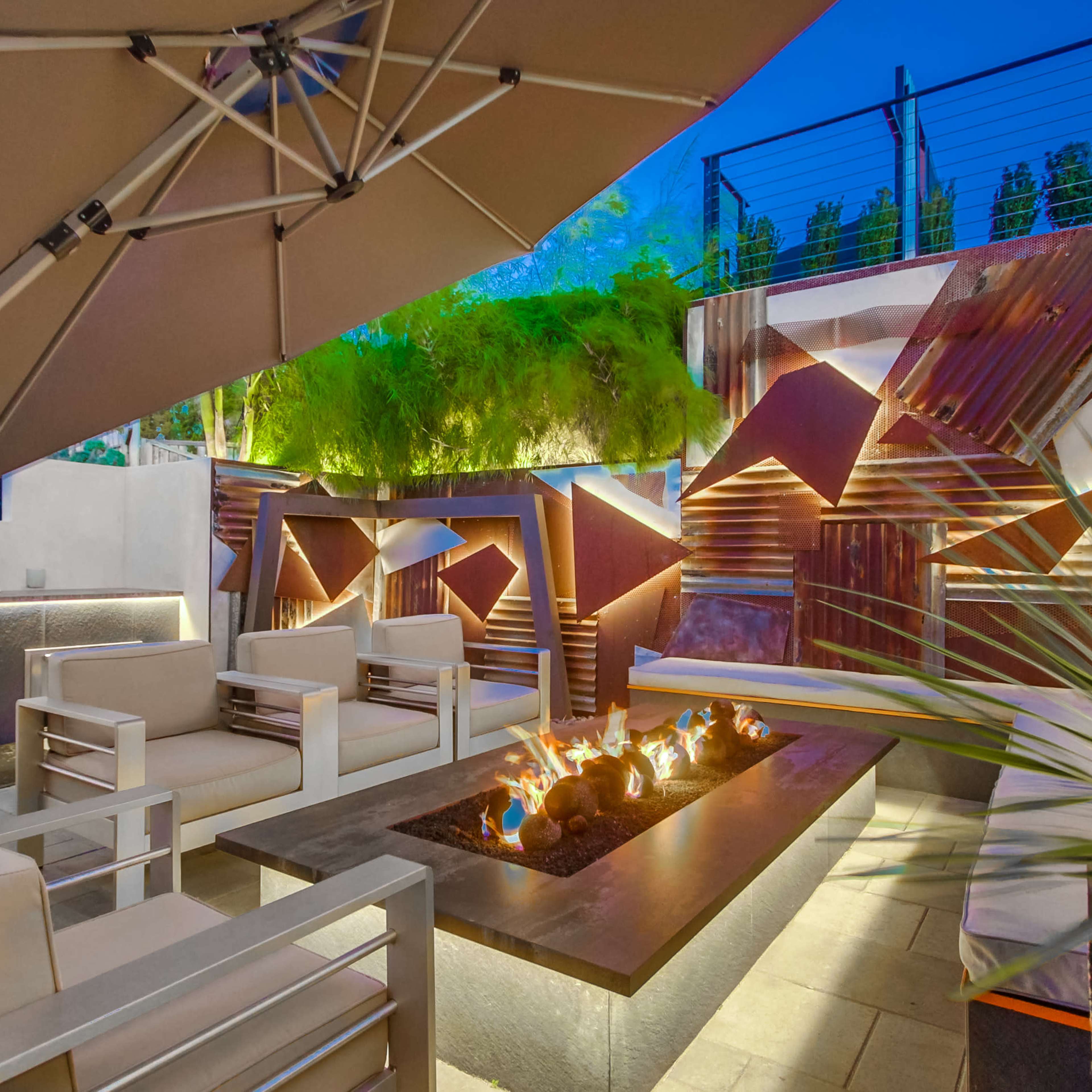 A modern outdoor lounge area with a fire pit, seating, and a barbecue grill, surrounded by greenery and illuminated architectural elements.