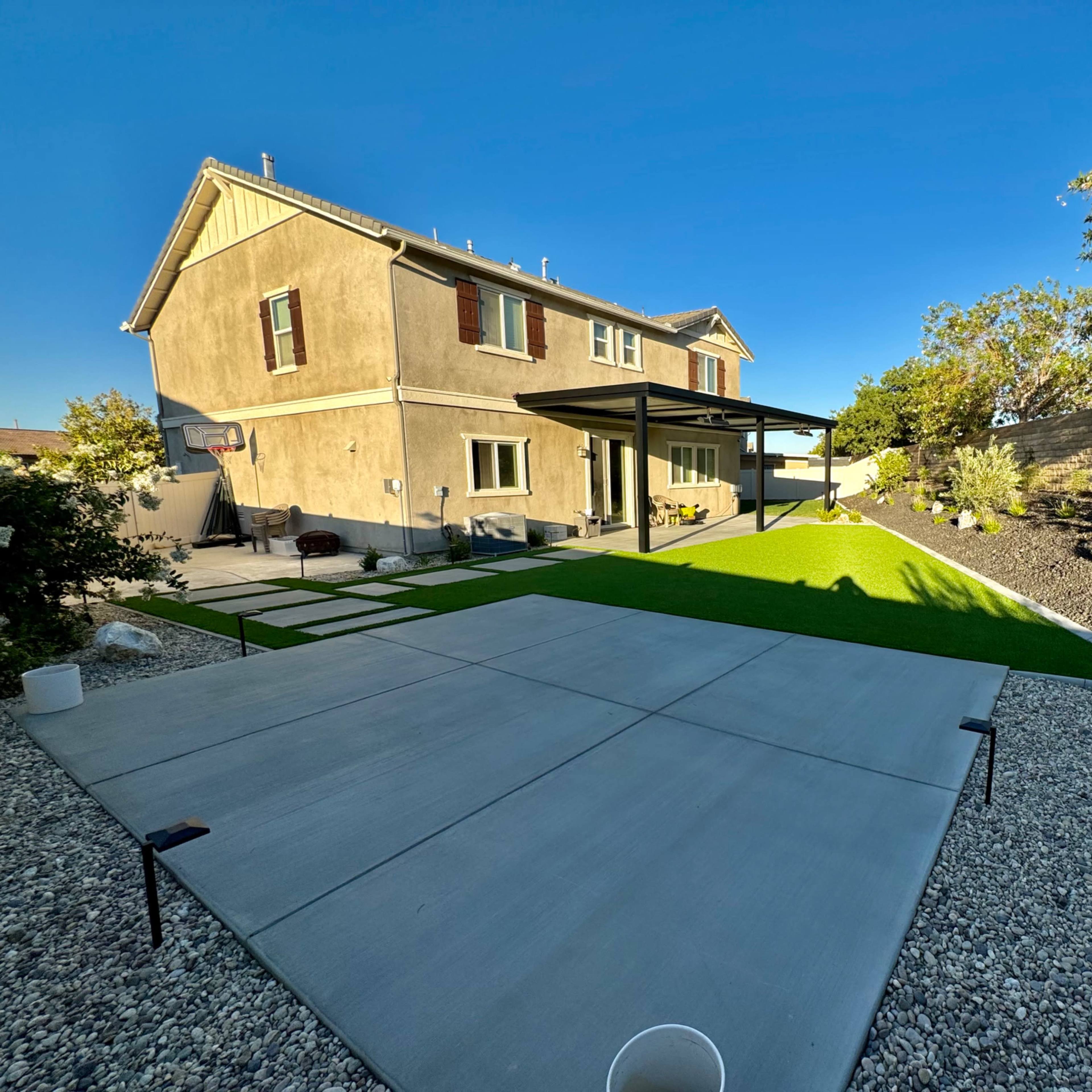 A backyard with a concrete patio, artificial grass, and a two-story house with a covered porch.