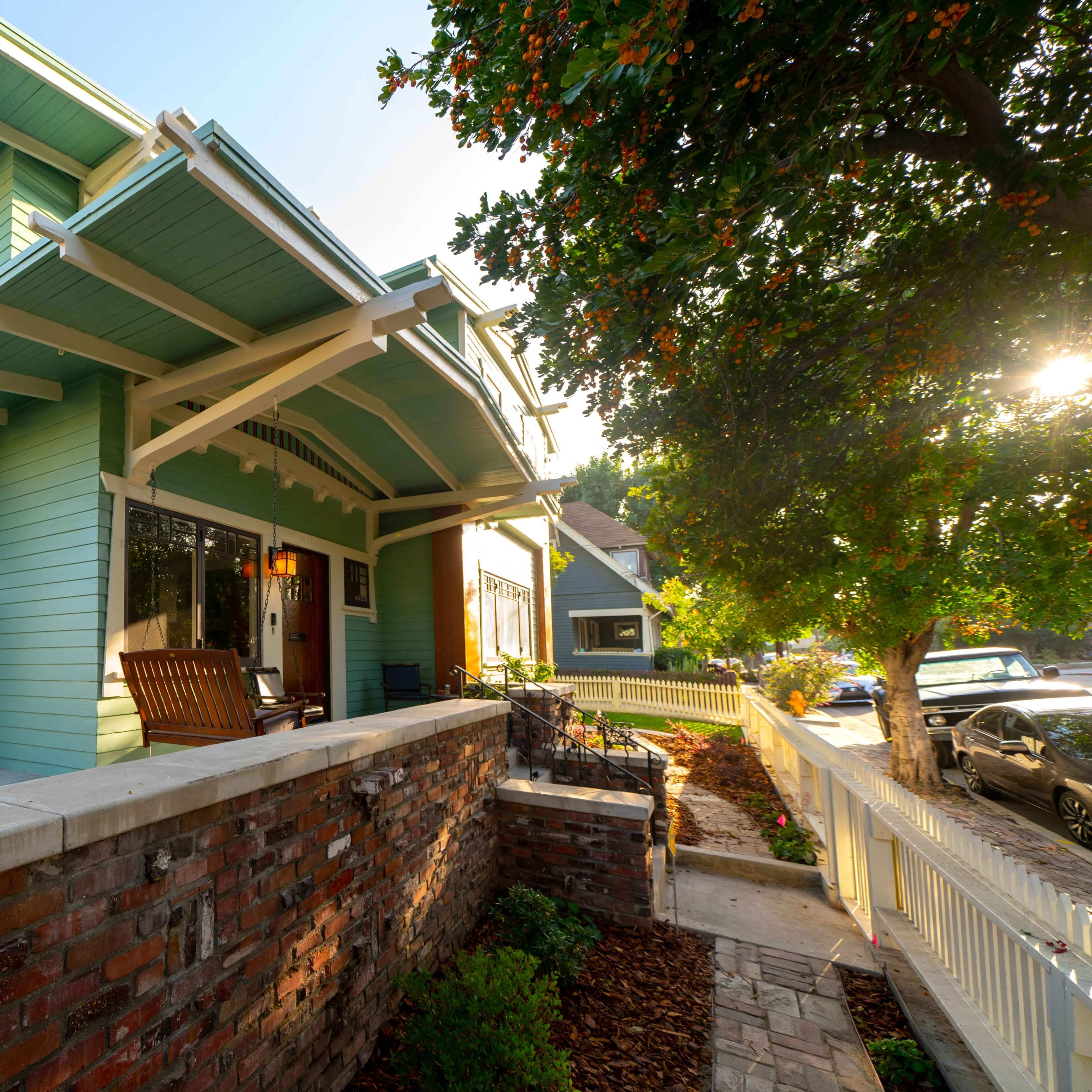 A blue-green Craftsman-style house with a front porch and seating area is situated on a corner lot, surrounded by landscaped gardens and parked cars.