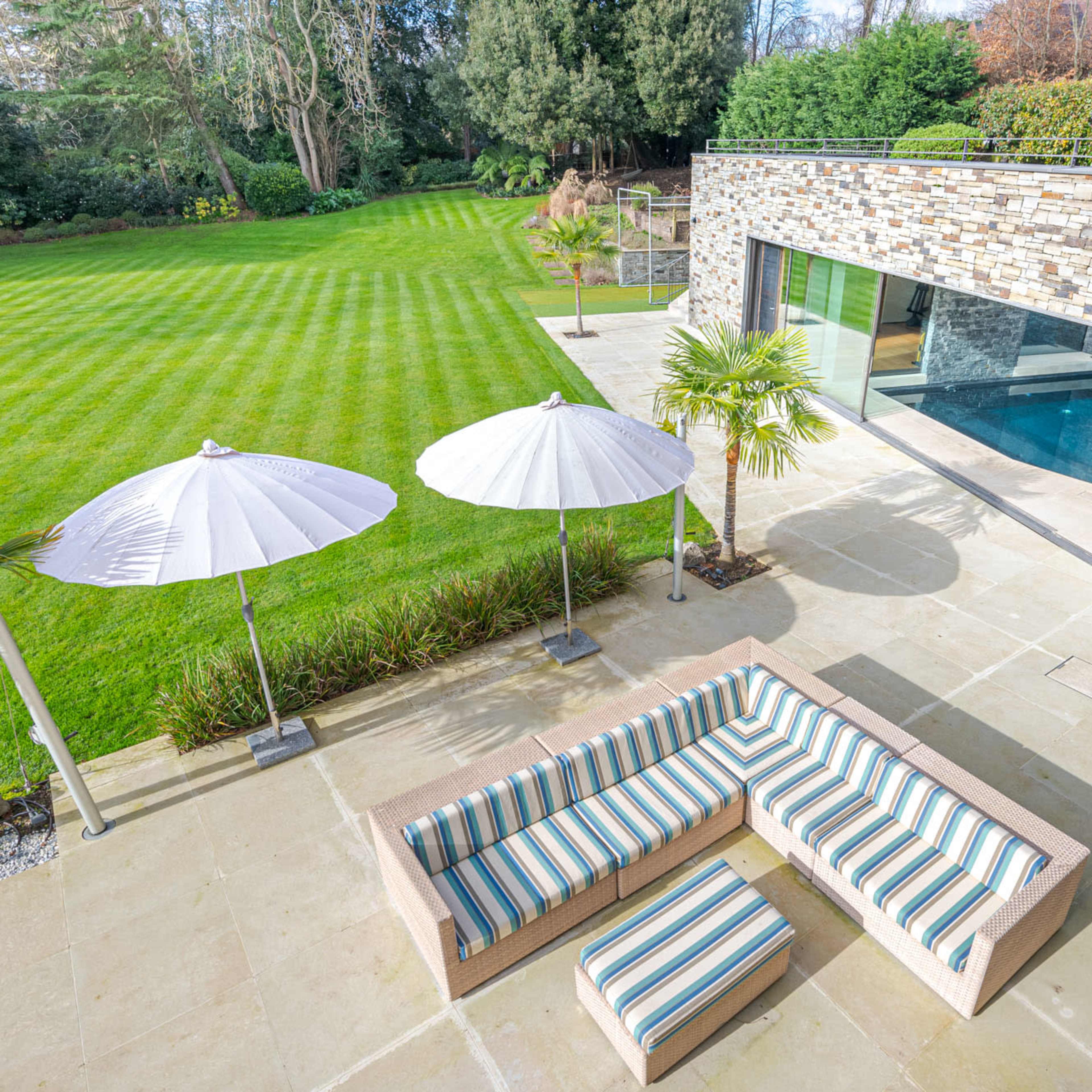 A modern outdoor patio area with striped seating, white umbrellas, and a pool adjacent to a landscaped lawn.