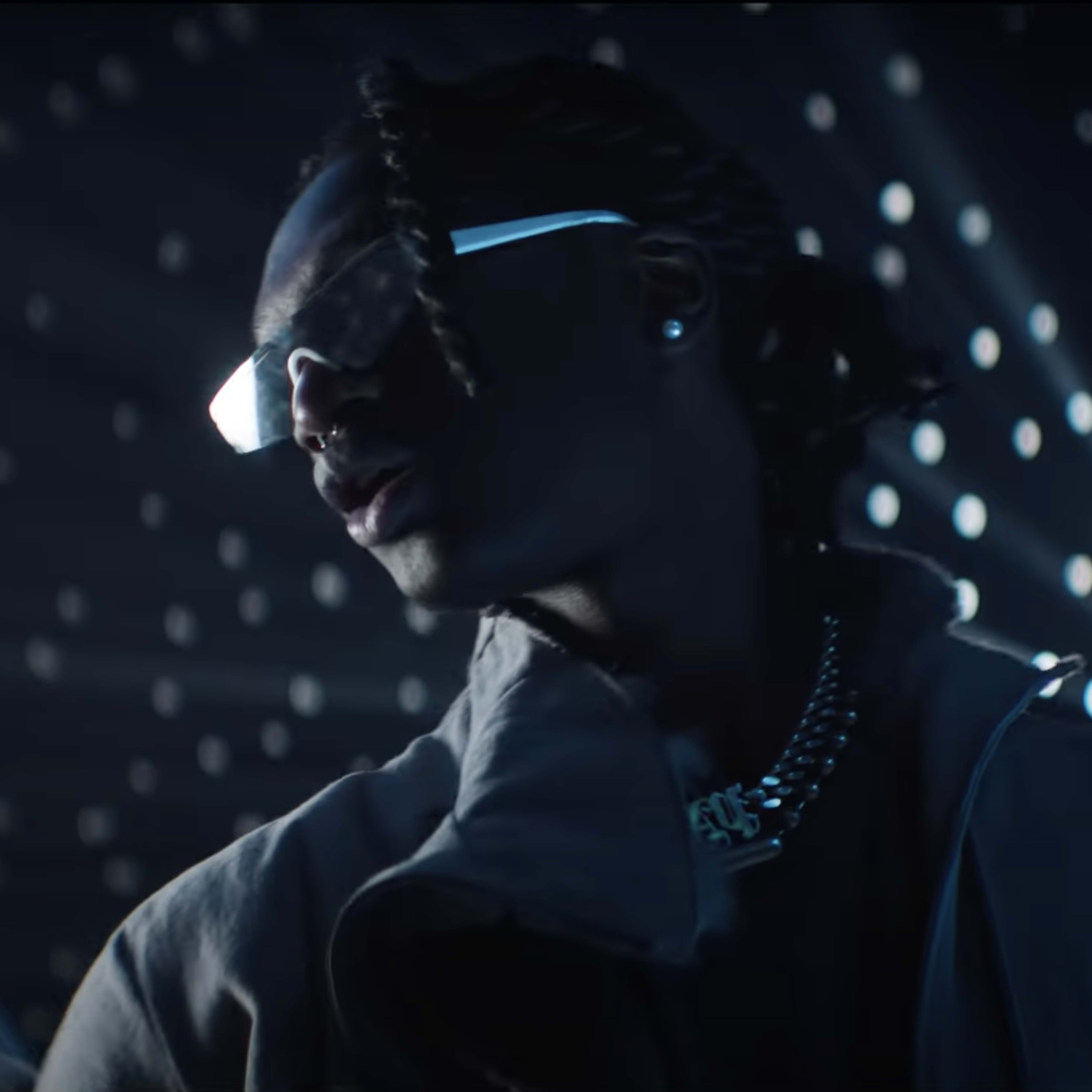A person wearing sunglasses and a chain stands in a dimly lit space with bright lights radiating around them.
