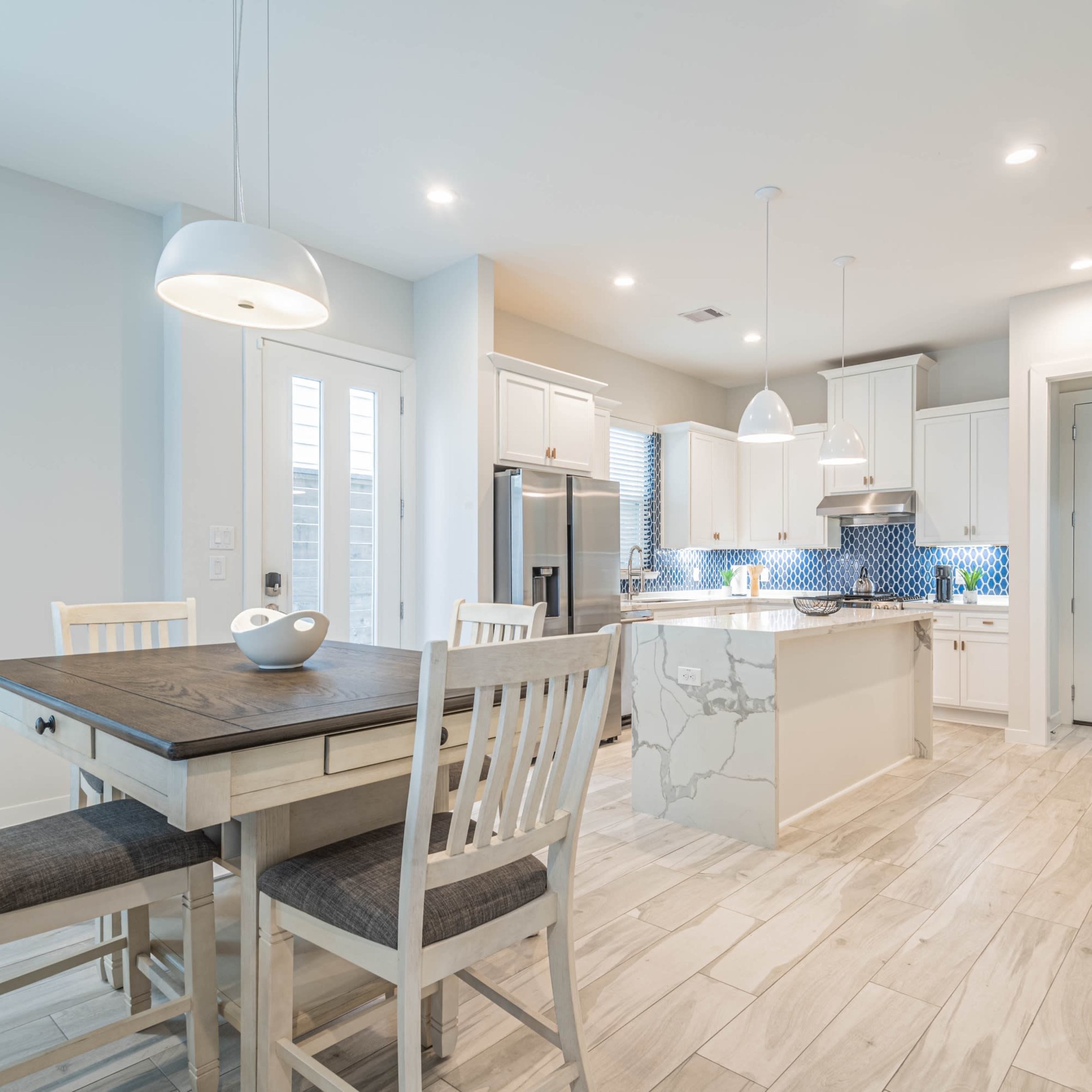 A modern kitchen and dining area feature a wooden table with white chairs, quartz countertops, stainless steel appliances, and blue tile backsplash.