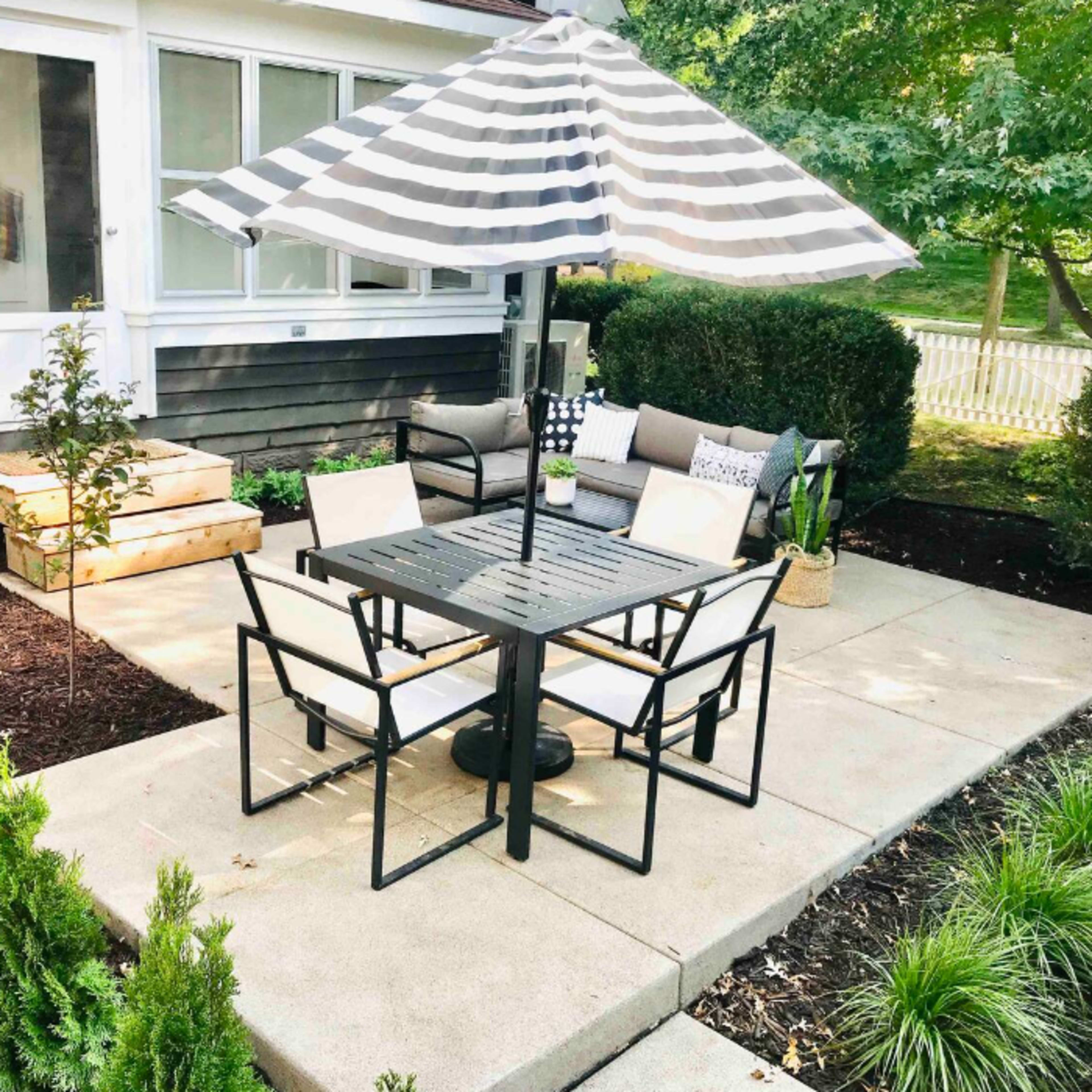 A patio area features a black striped umbrella over a table with four chairs, accompanied by a couch and greenery surrounding the space.