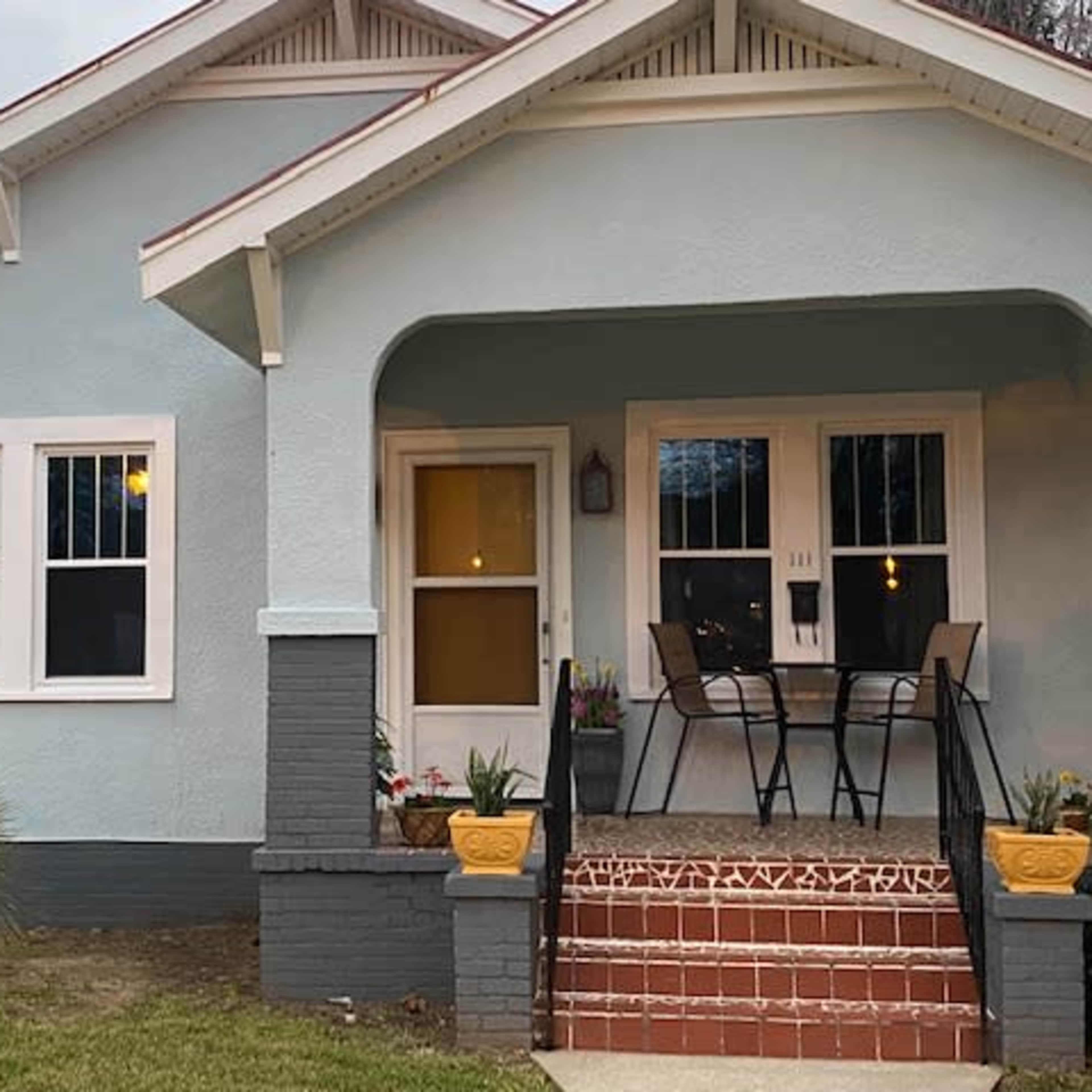 A light blue house with a covered front porch features two chairs and a small table, surrounded by well-maintained landscaping.