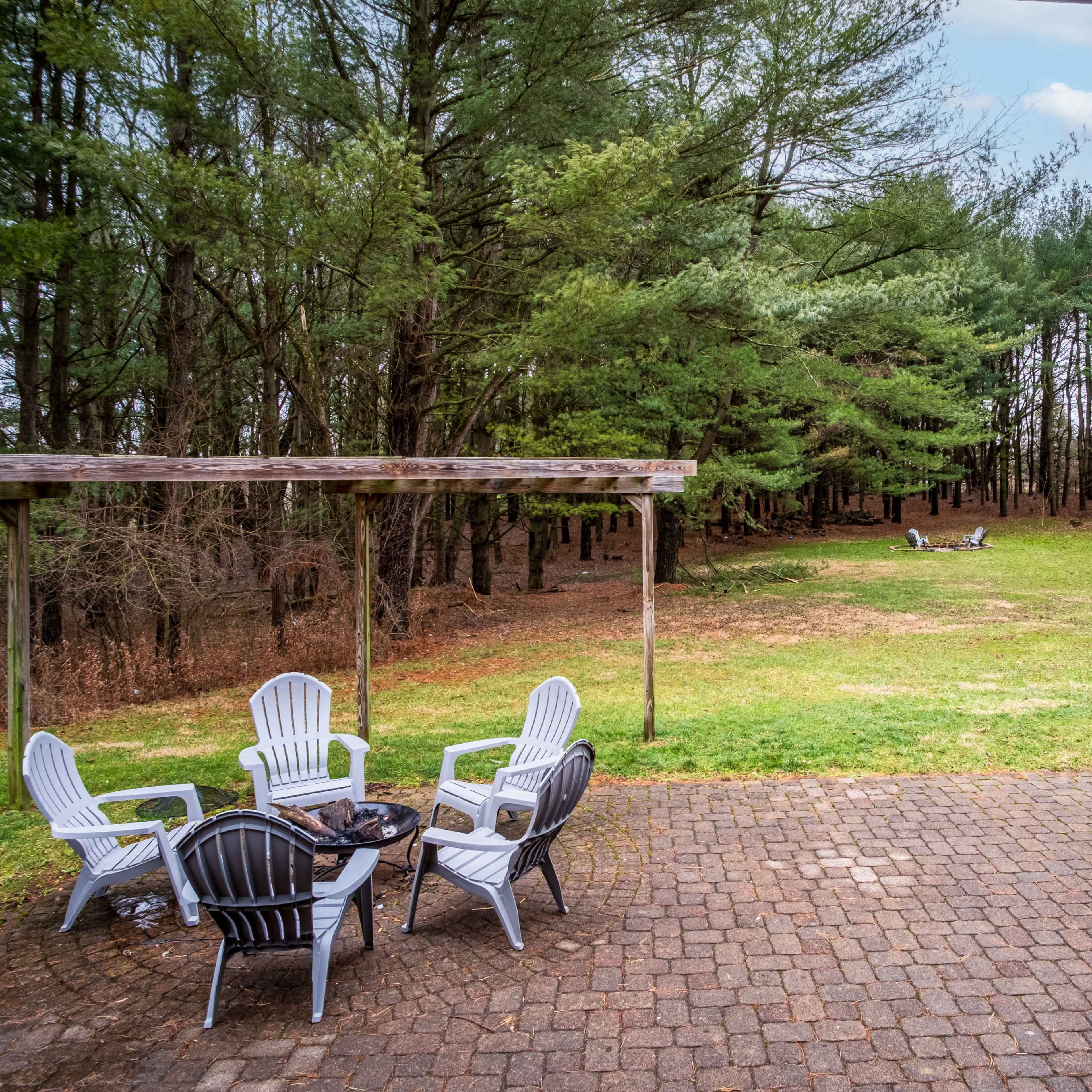 A stone patio with white lawn chairs overlooks a grassy area surrounded by pine trees.