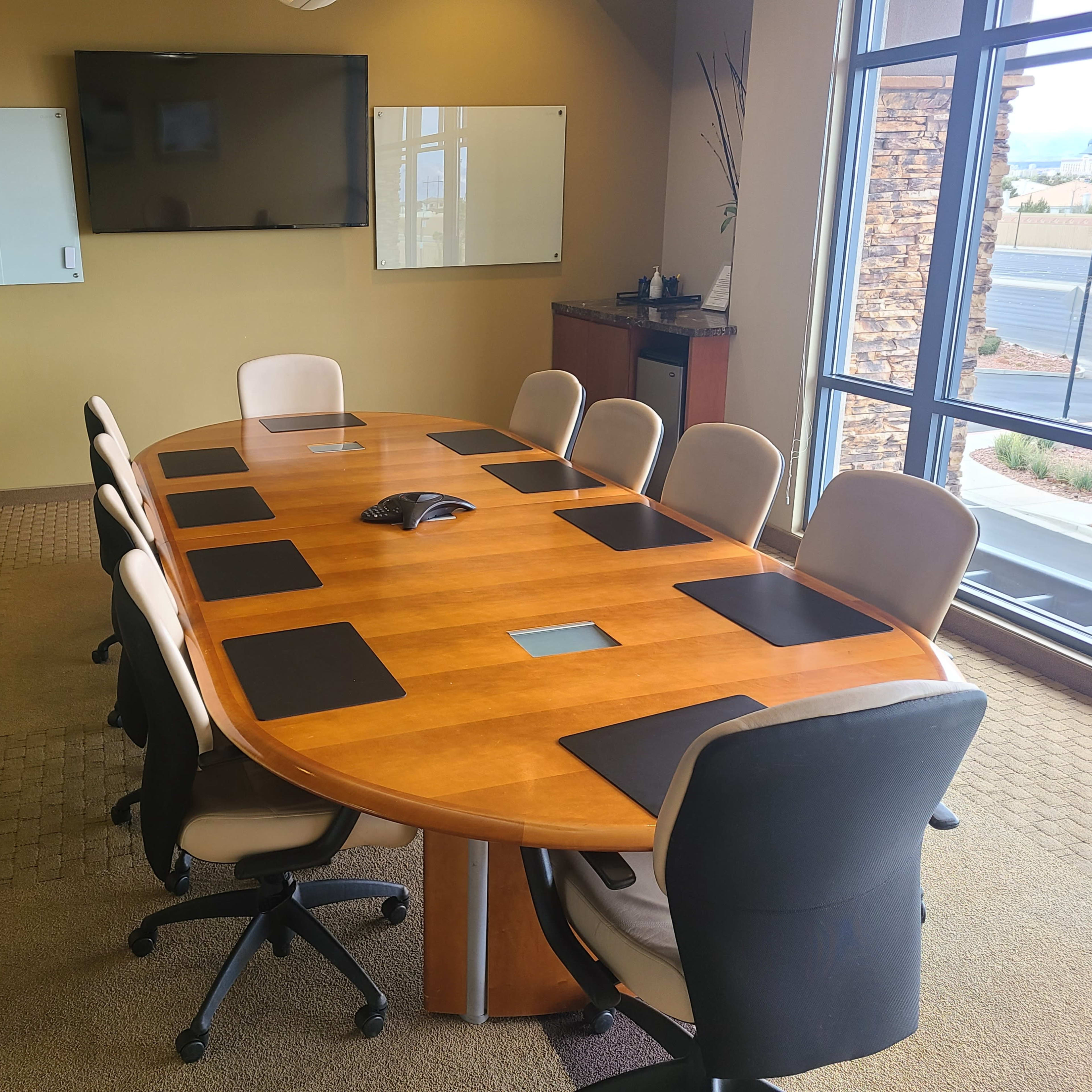 A conference room features a large oval wooden table surrounded by several office chairs, with a wall-mounted screen and whiteboards in the background.