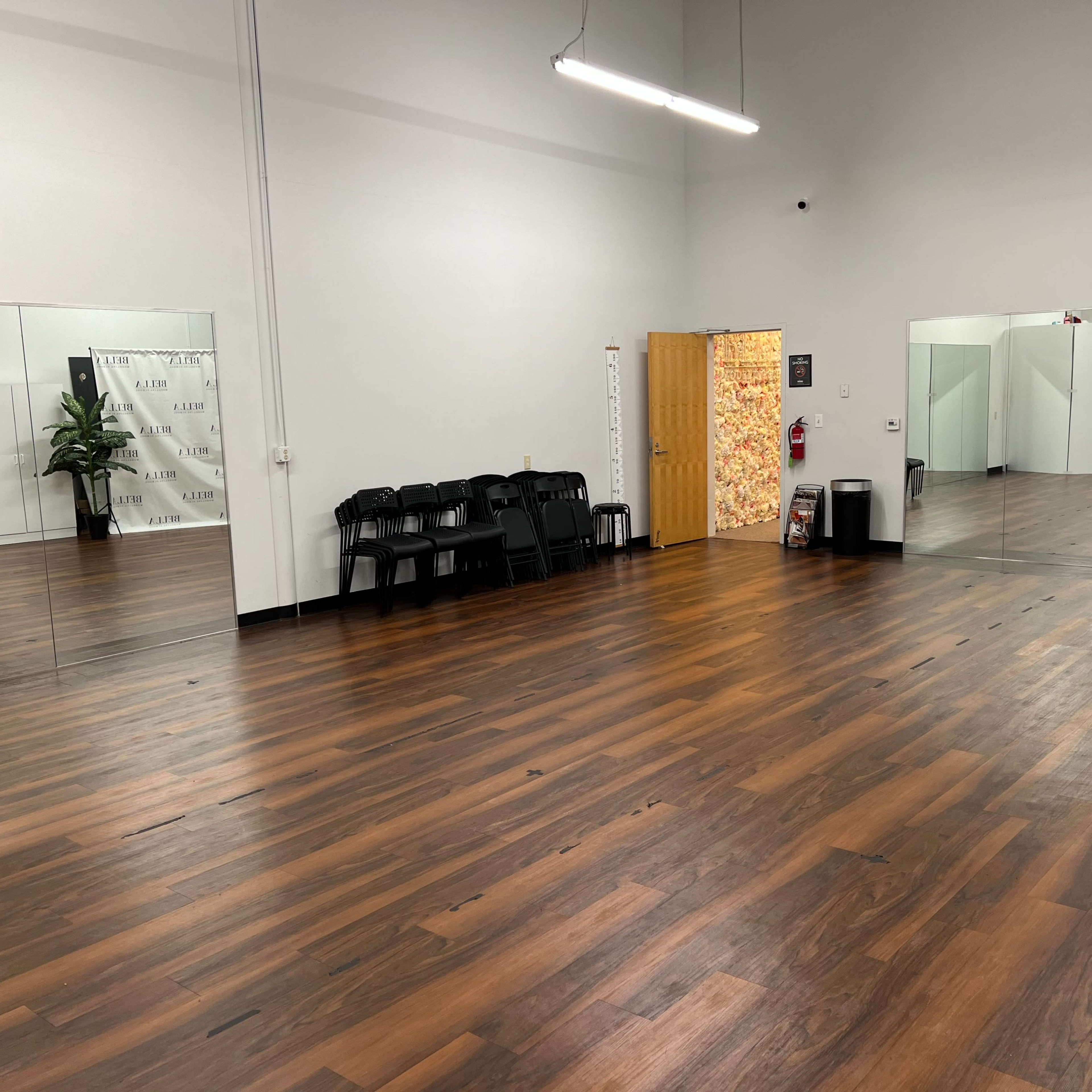 A spacious room with hardwood flooring, several black chairs stacked neatly against one wall, large mirrors on one side, and a colorful door at the end of the room.
