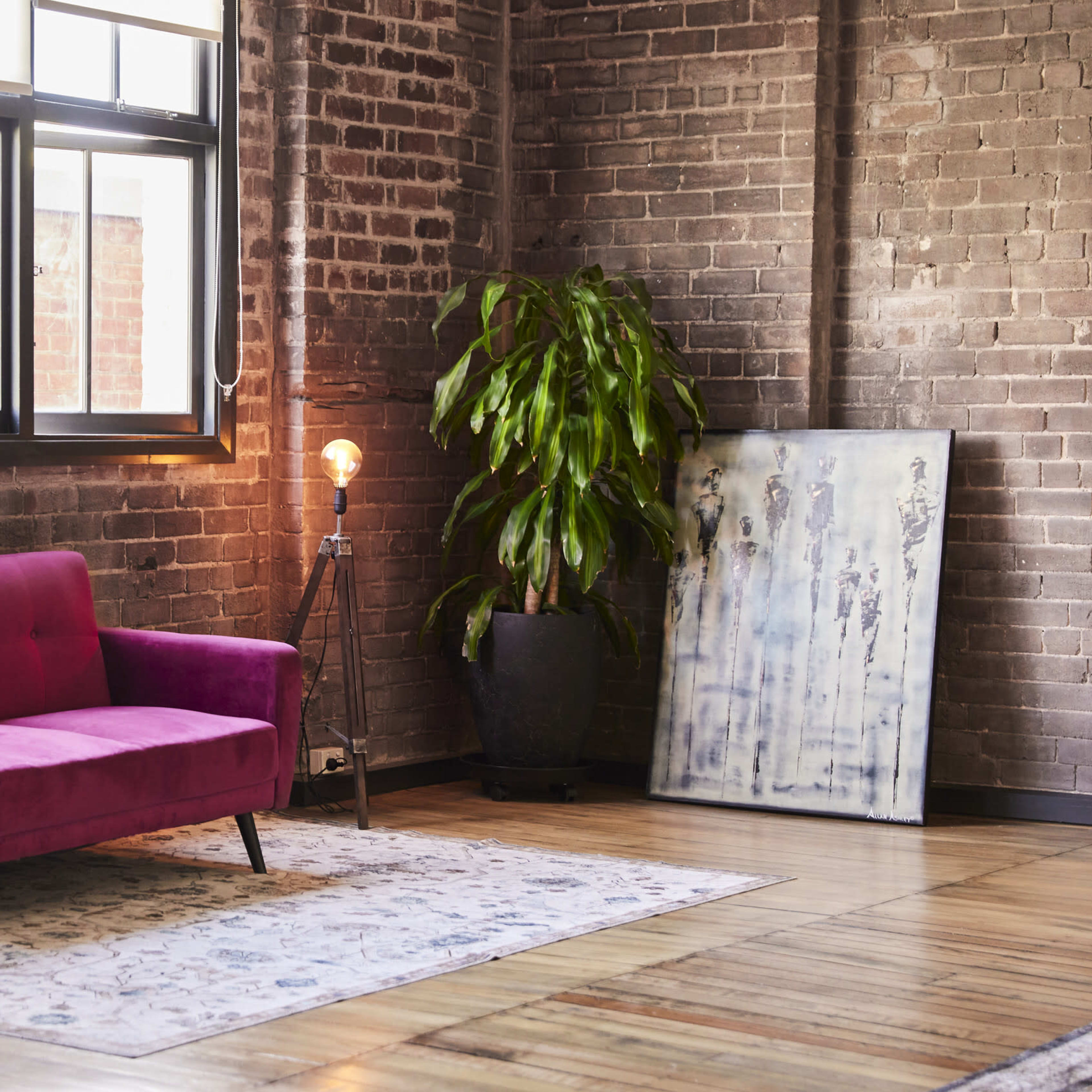 A room with a pink sofa, a blue chair, a large mirror, a plant, and a piece of art against a brick wall.