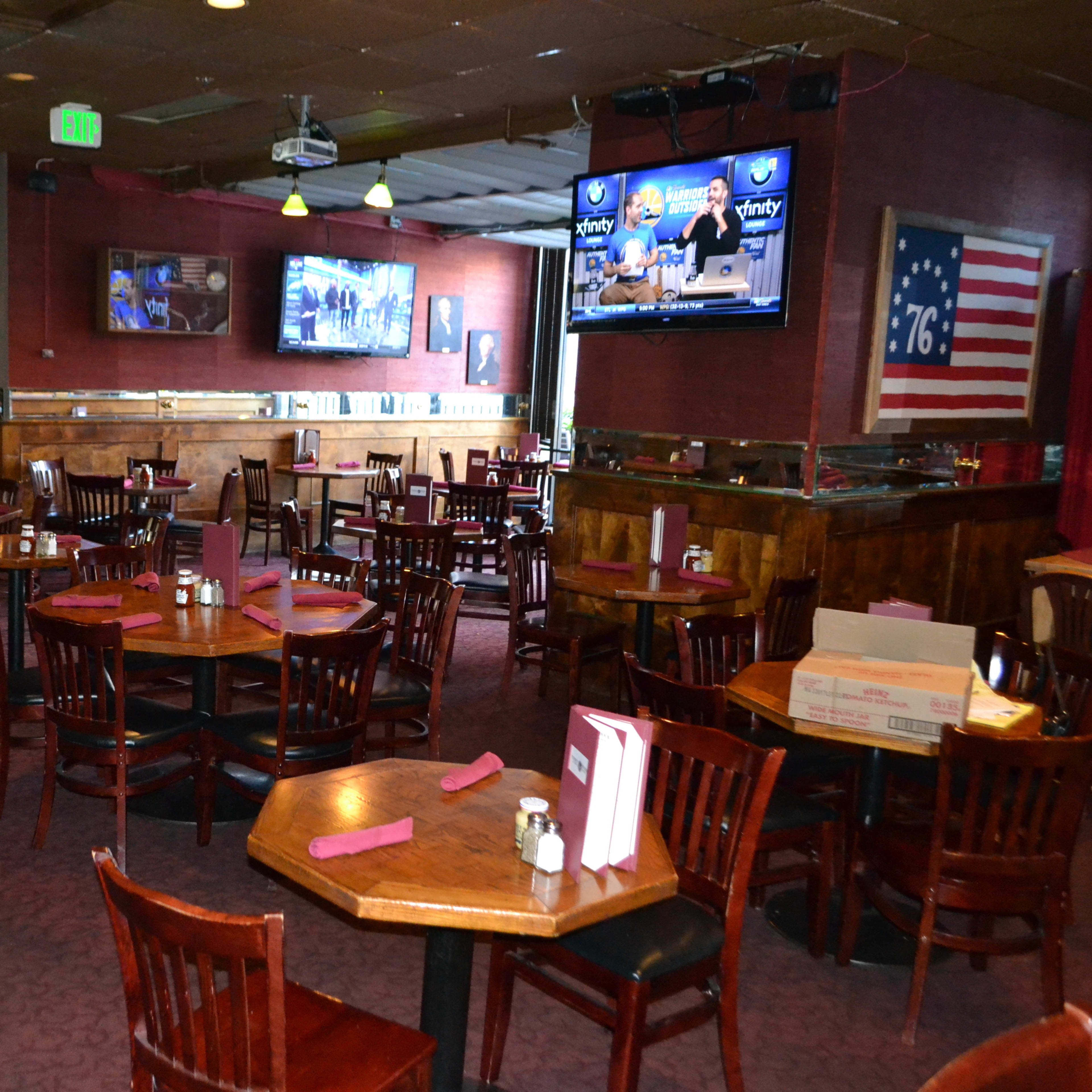 A mostly empty restaurant with wooden tables and chairs, two televisions mounted on the walls, and a large American flag displayed.