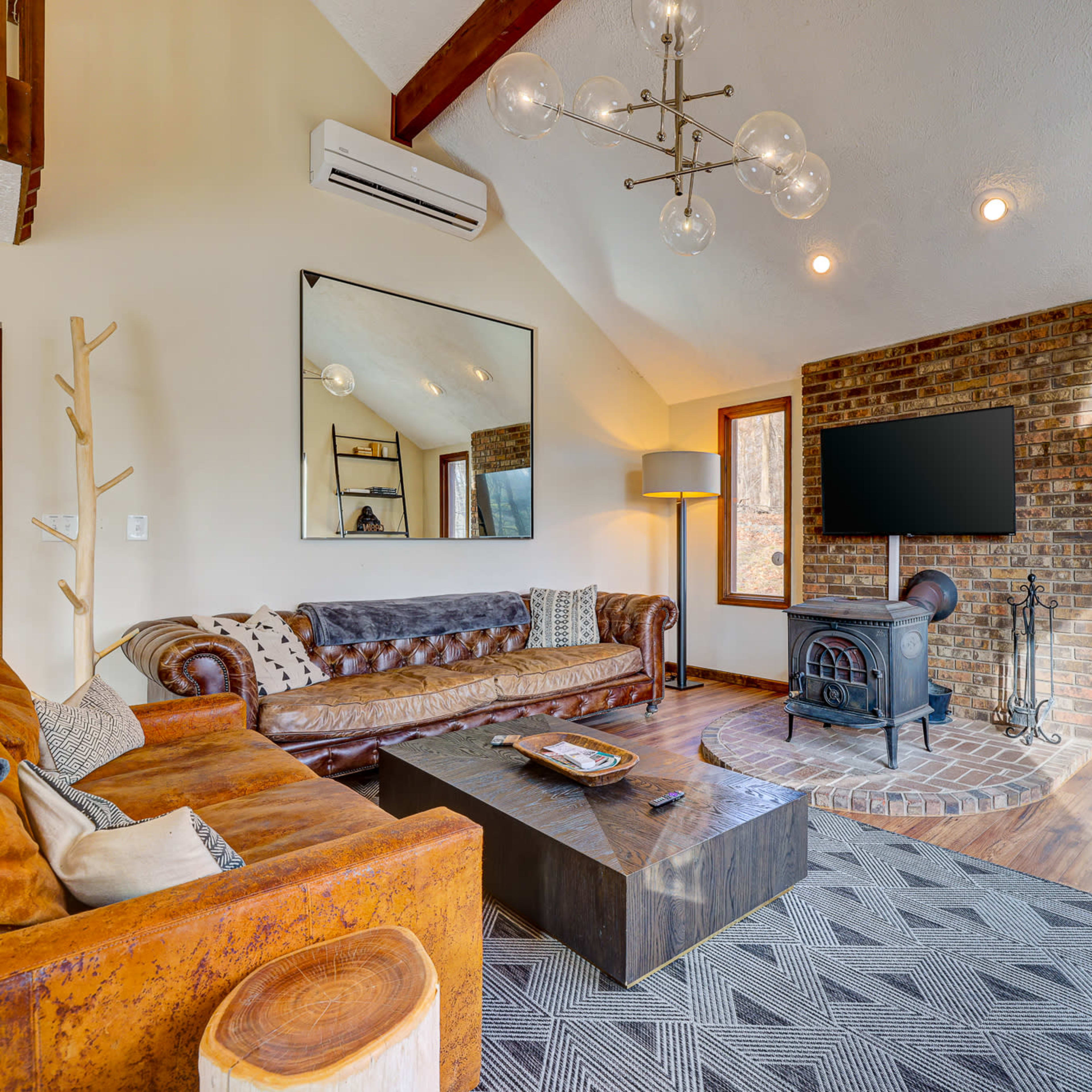 A modern living room features a leather sofa, a wooden coffee table, a brick fireplace, and large windows overlooking a wooded area.