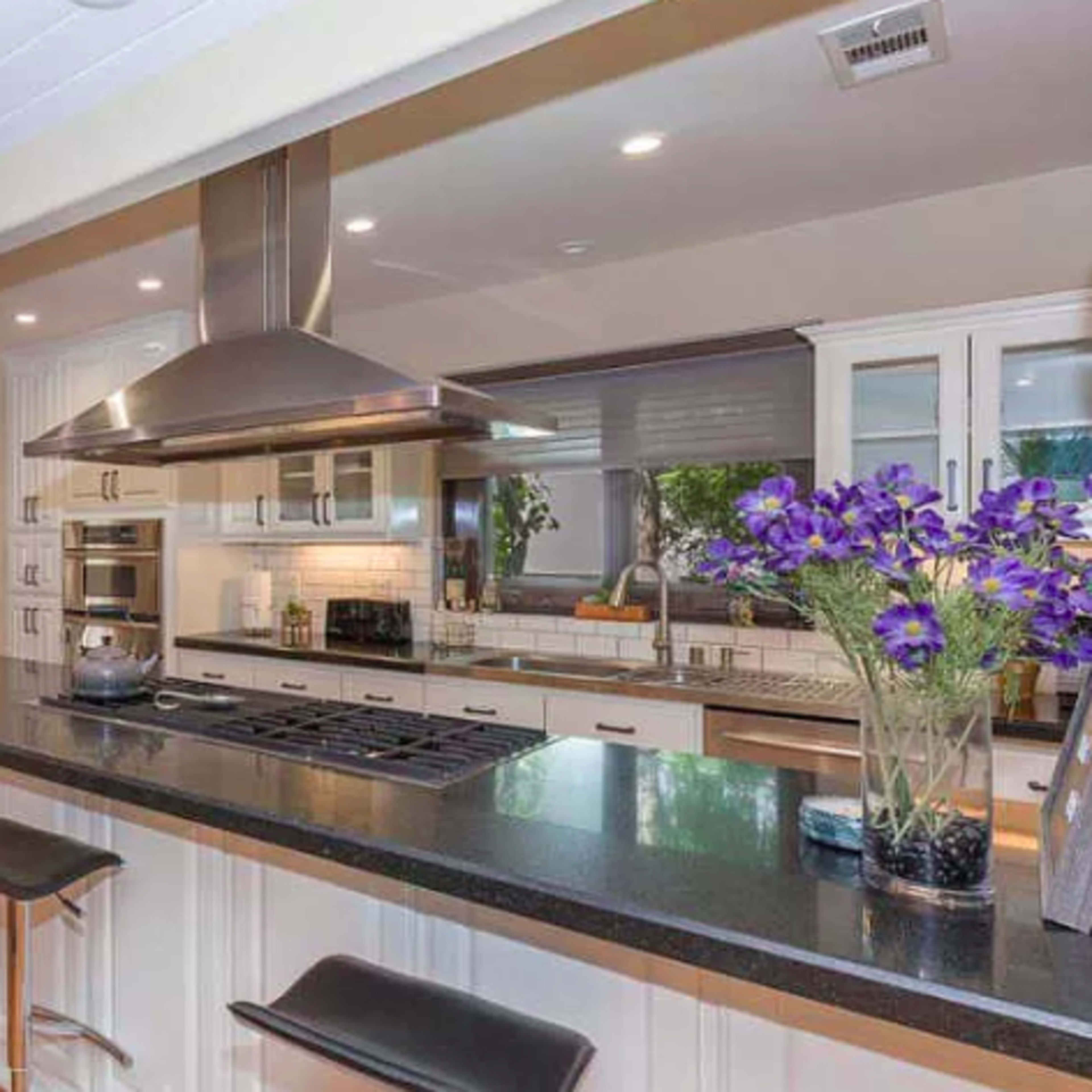 A modern kitchen with a large island, stainless steel appliances, and bar stools, featuring wooden flooring and natural light.