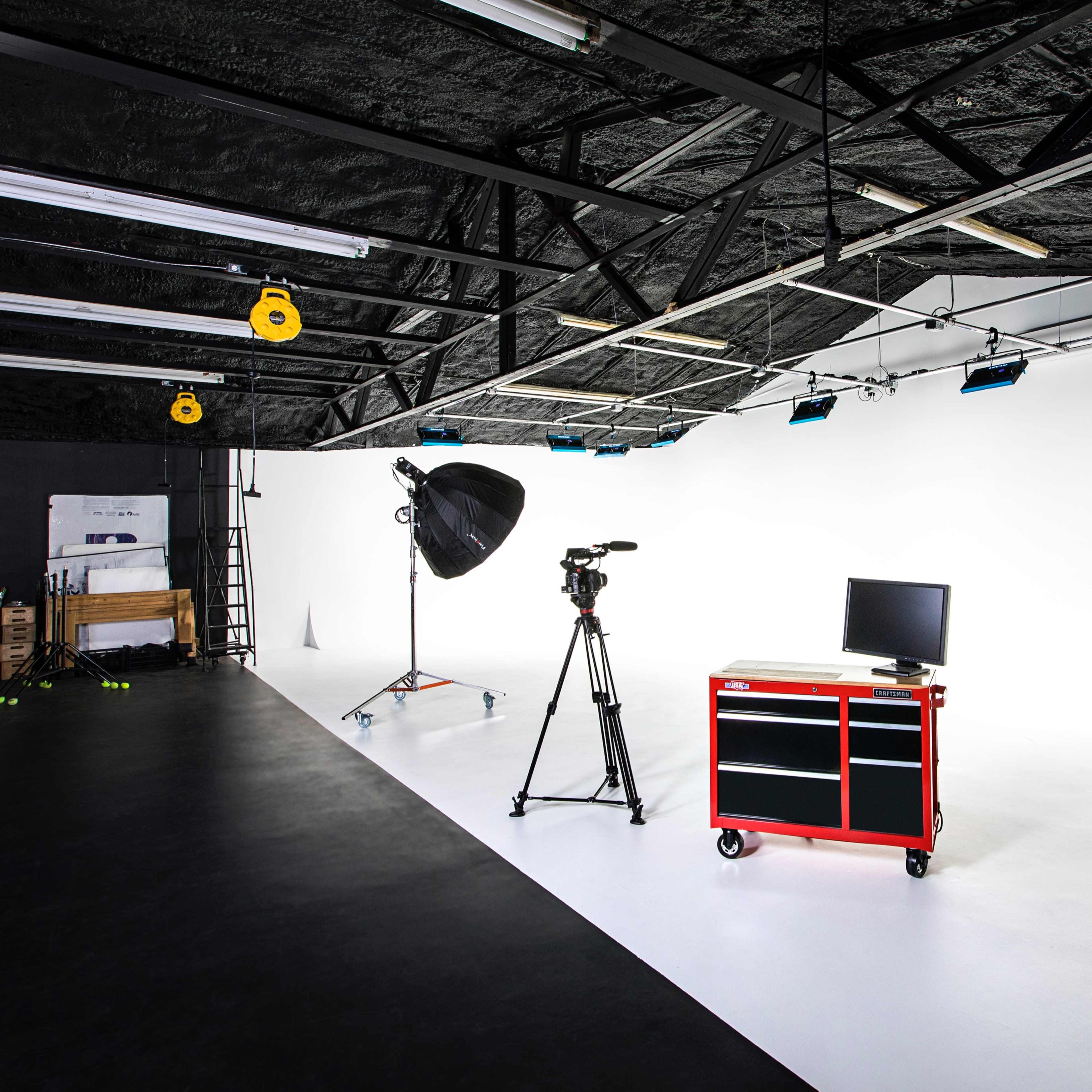 A photography studio with a camera on a tripod, a large light umbrella, a red tool cart, and a monitor against a white backdrop.