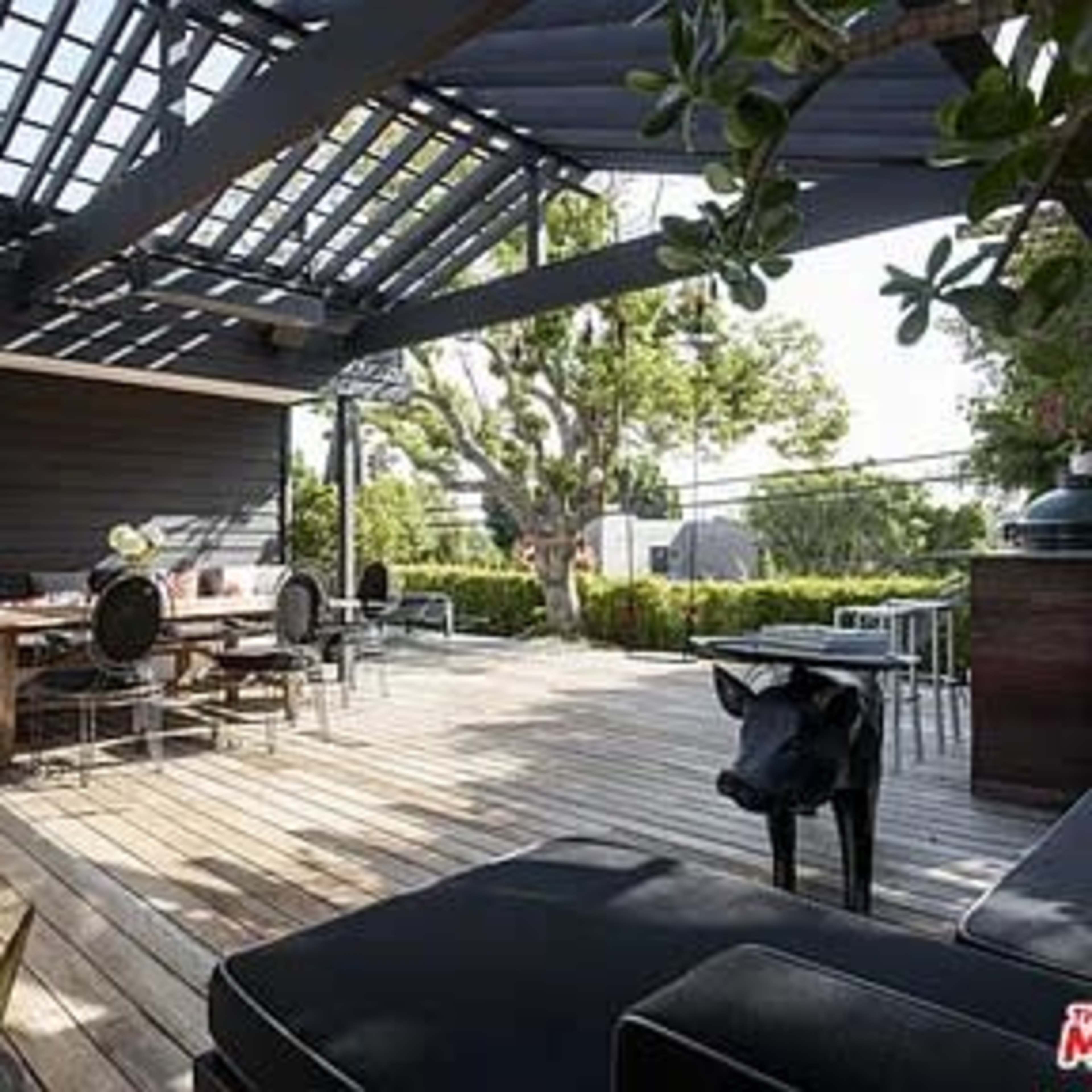 A wooden-decked outdoor living space with a dining area, seating, and a trellis overhead, surrounded by greenery.