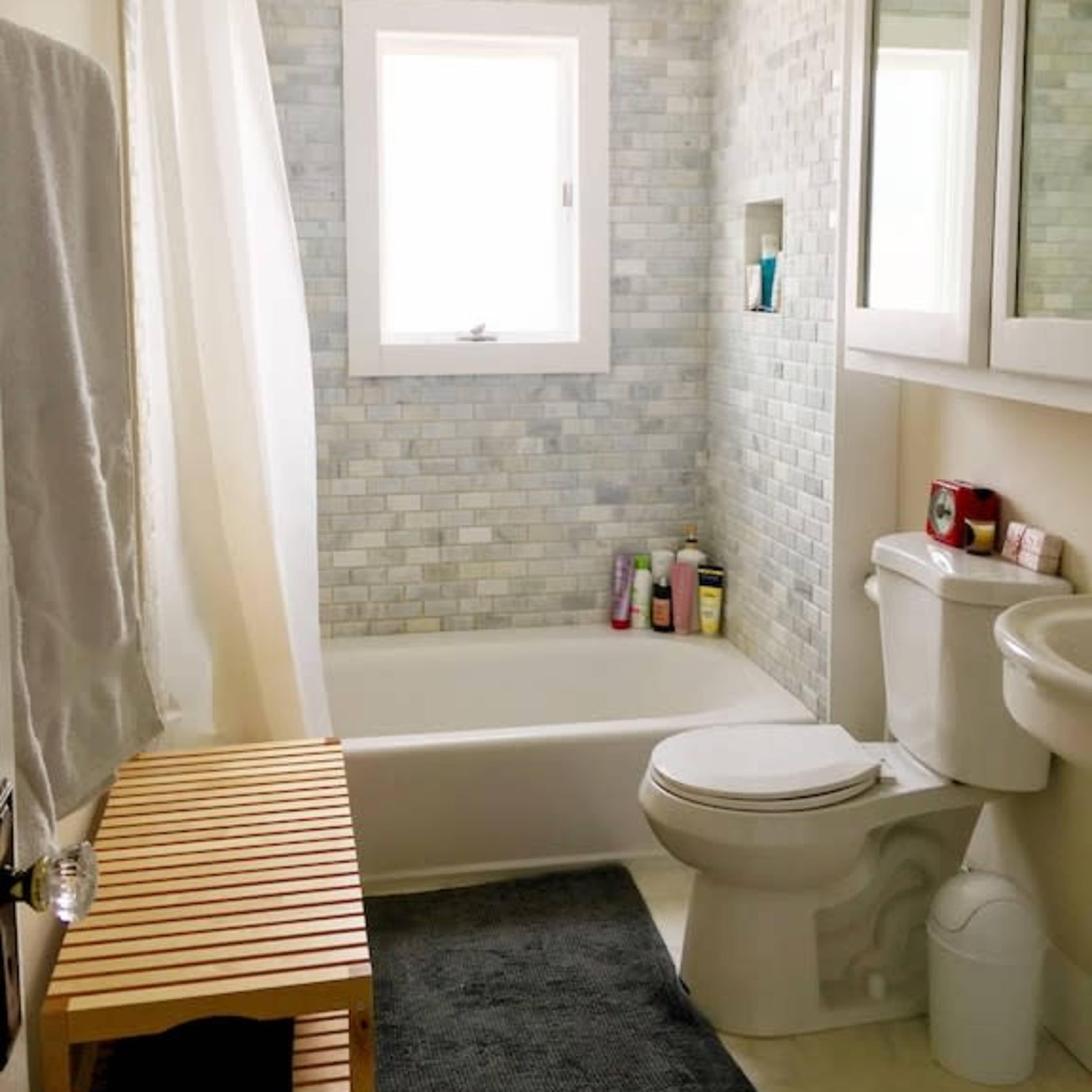 A modern bathroom featuring a bathtub with a shower curtain, a tiled wall, a small wooden bench, a toilet, and a mirror cabinet above the sink.