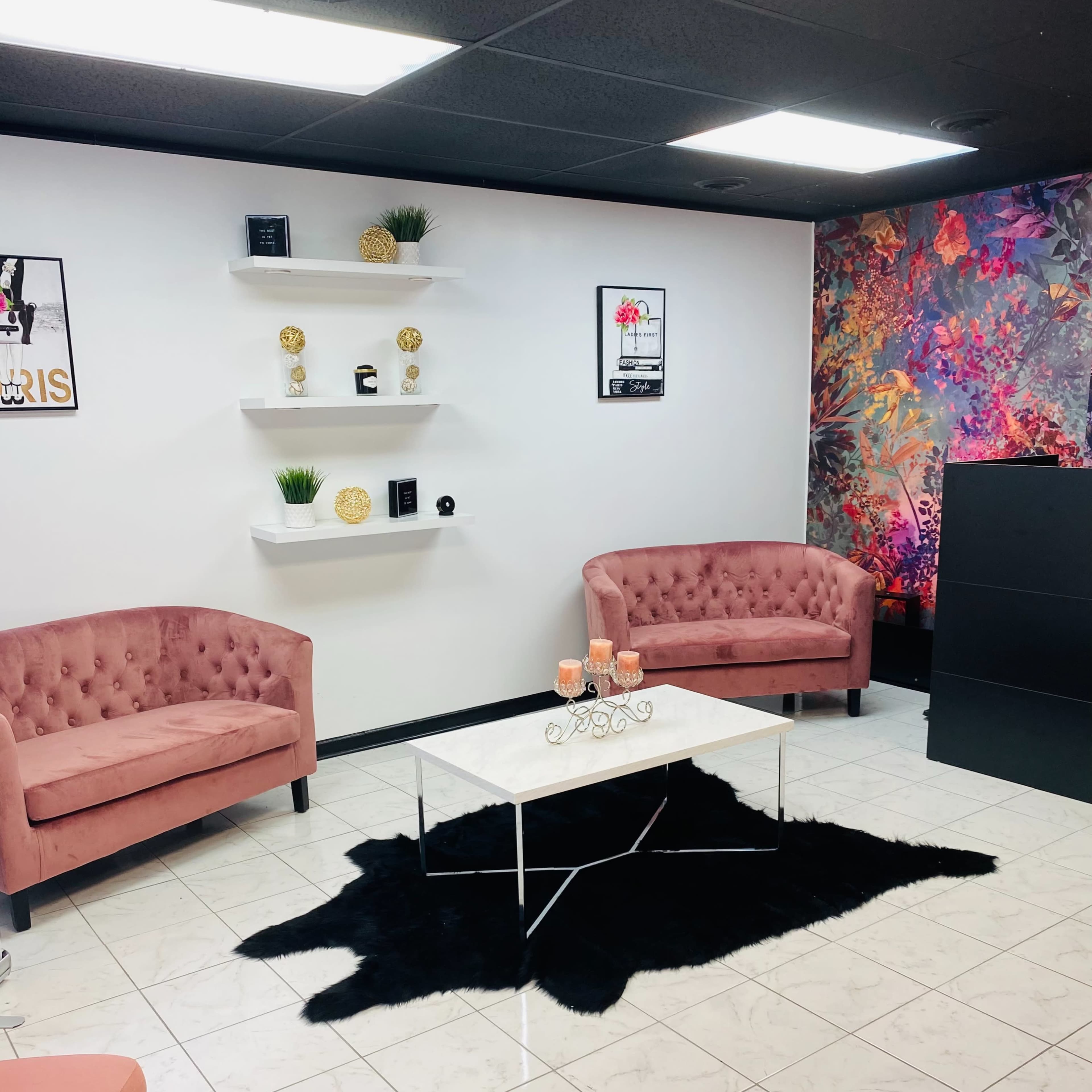 A modern waiting area with two pink tufted chairs, a white coffee table on a black rug, and decorative shelves on a white wall.