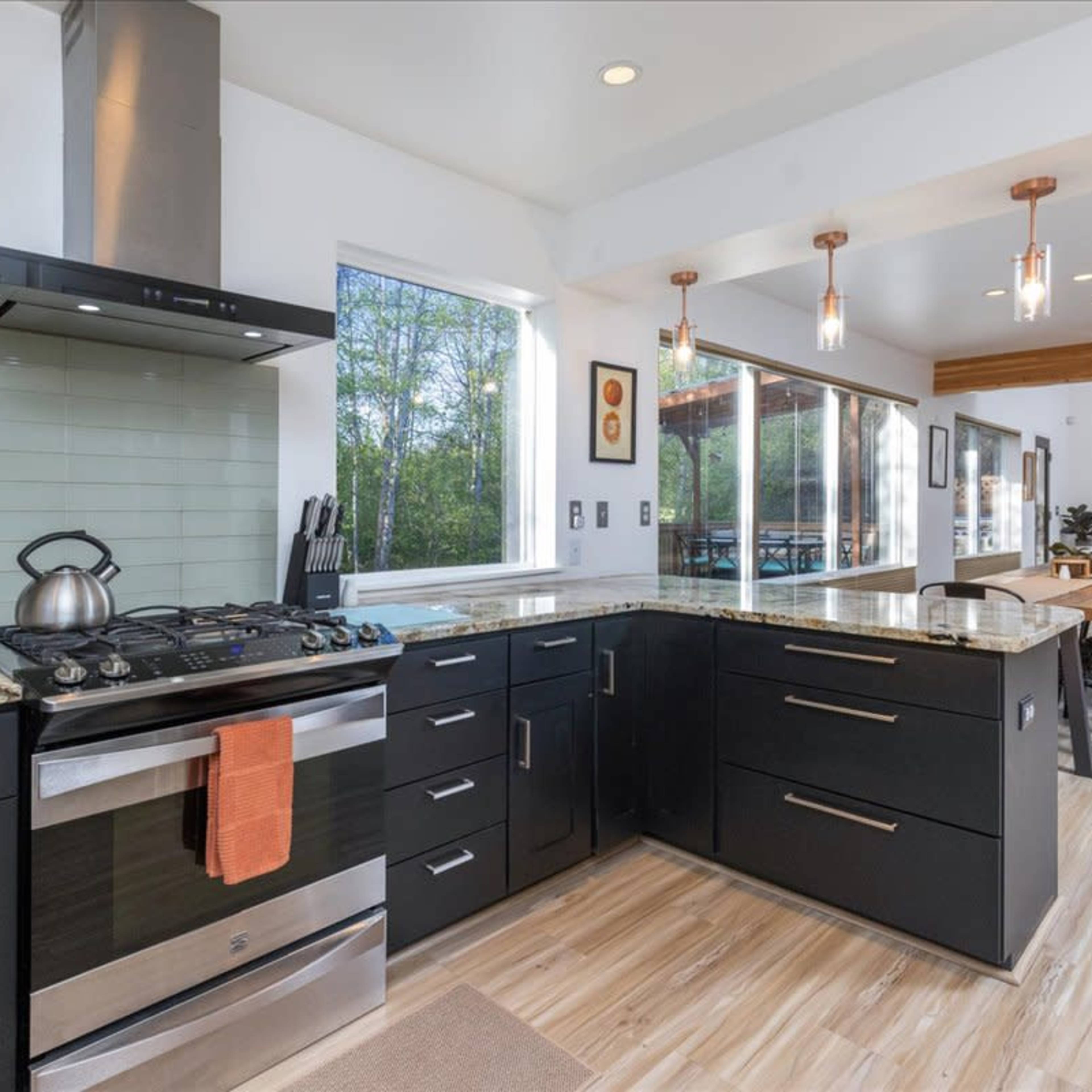 A modern kitchen with black cabinets, a stainless steel stove, and a granite countertop, adjacent to a dining area with pendant lighting.