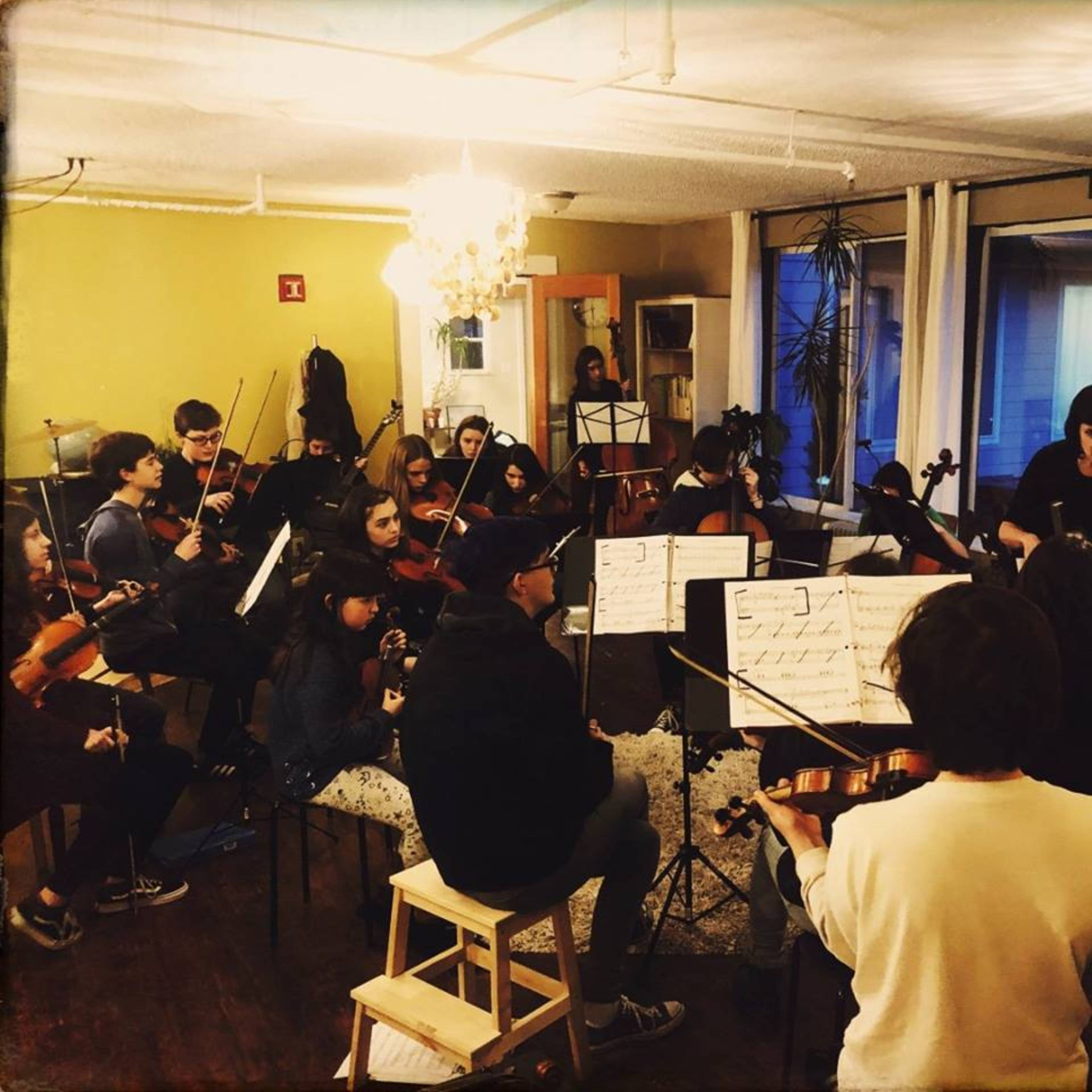 A group of musicians is seated in a studio, playing string instruments while reading sheet music.