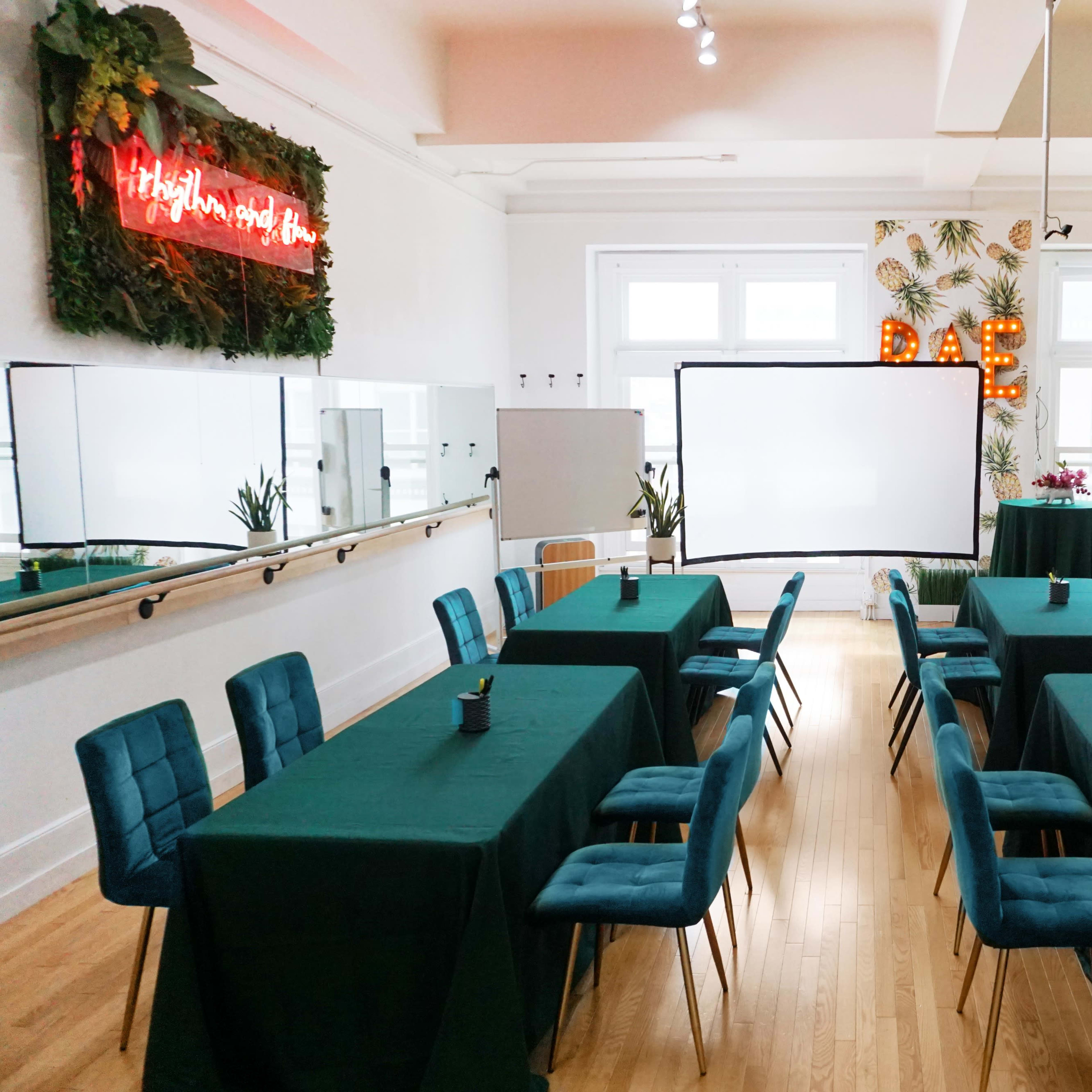 A room set up for a conference, featuring several tables covered with green cloths and chairs arranged around them, along with screens and decorative plants in the background.