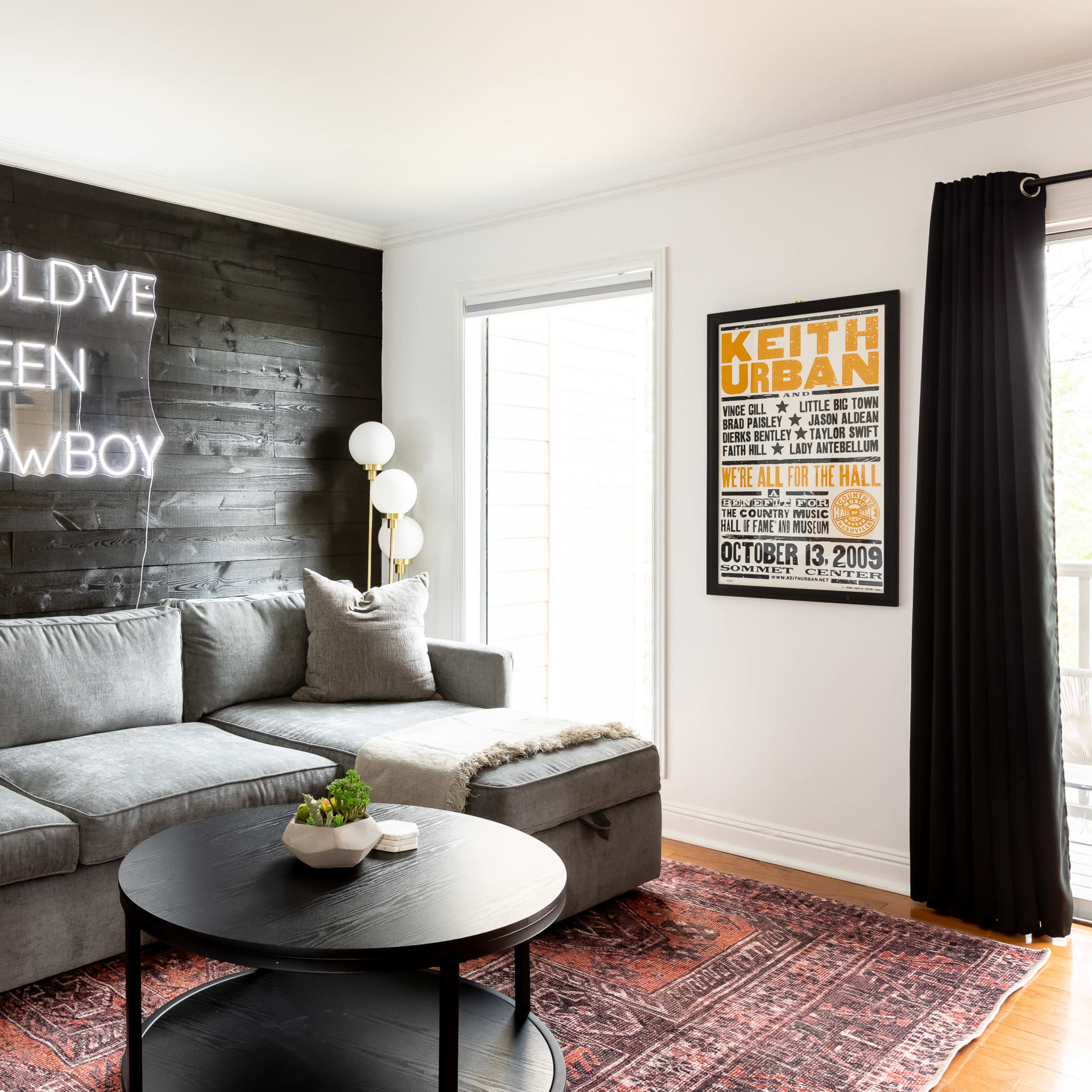 The living room features a gray sectional sofa, a round black coffee table, a neon sign on the wall that reads "SHOULD'VE BEEN A COWBOY," and a framed poster of a concert by Keith Urban.
