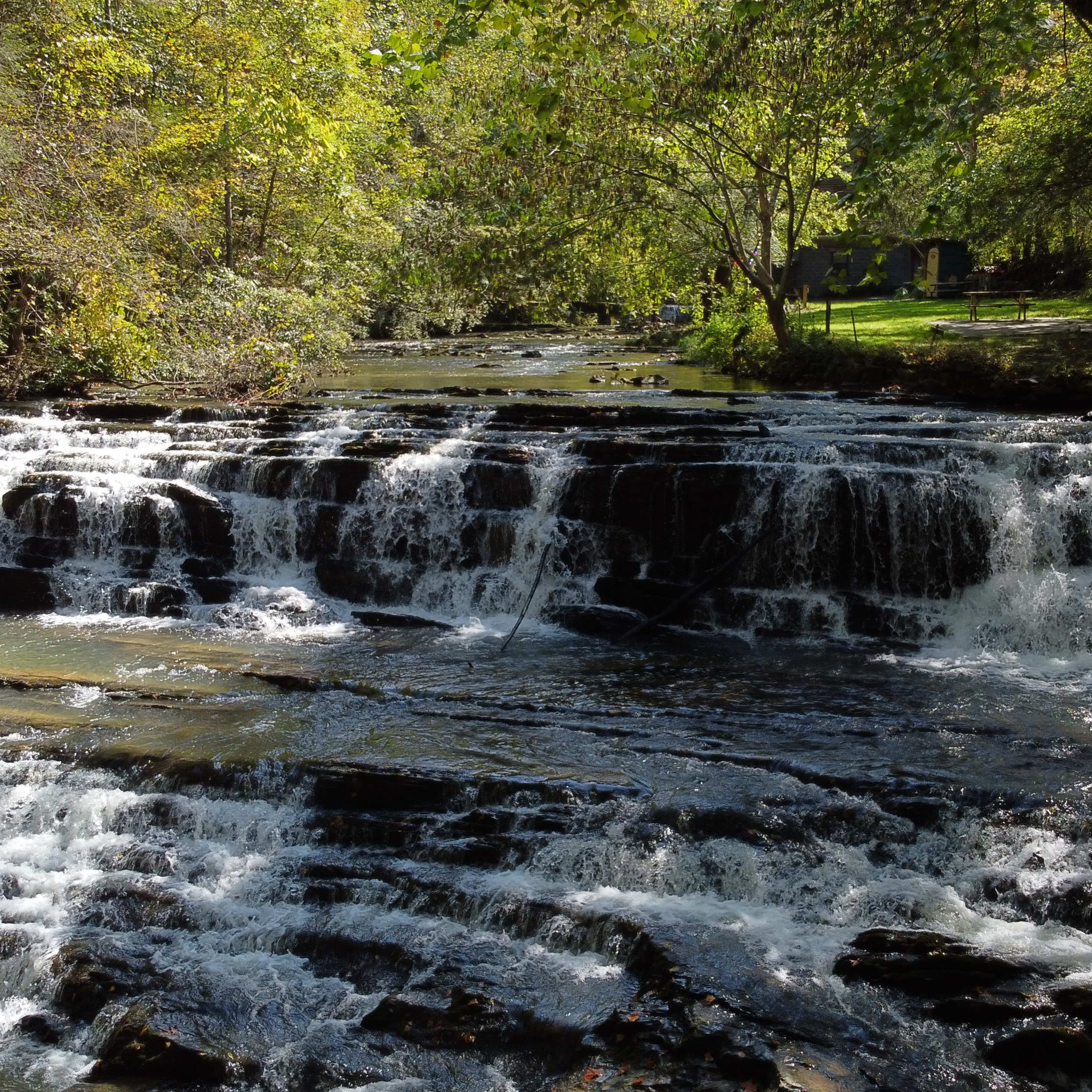 A waterfall cascades over rocky steps surrounded by lush greenery and trees.