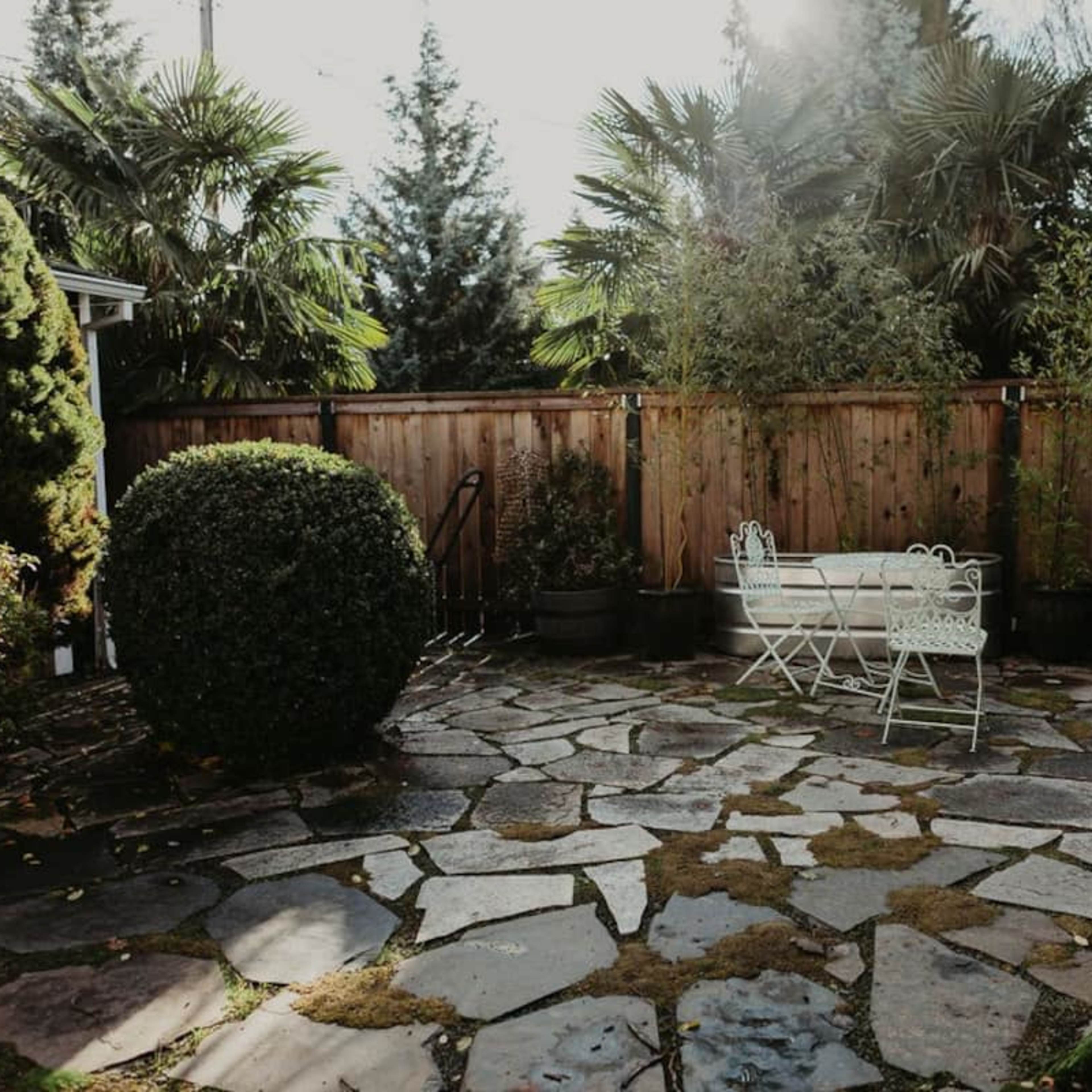 A stone patio surrounded by greenery, featuring a small white table and chairs in the center.