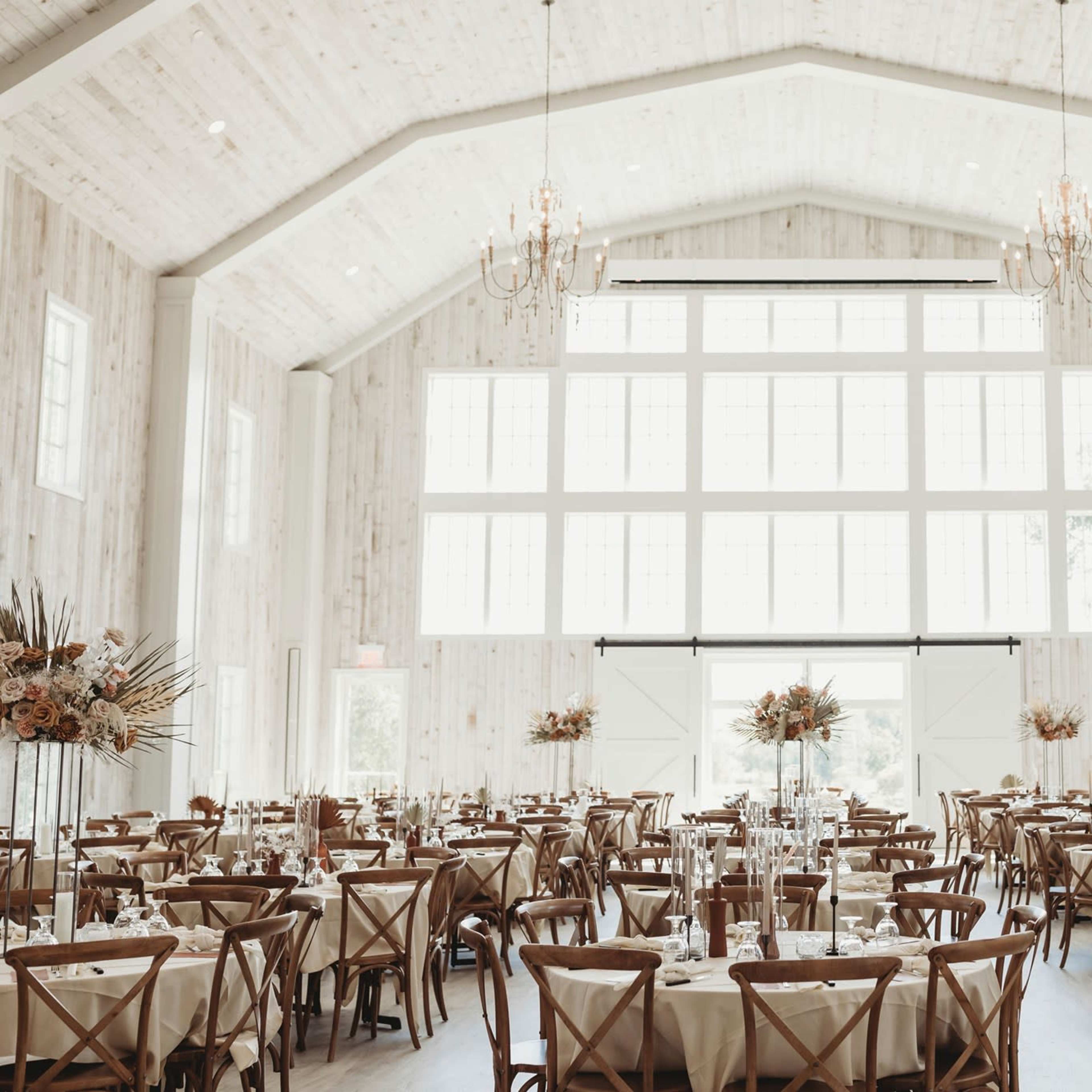 A large banquet hall with multiple wooden tables set for a formal event, featuring floral centerpieces and large windows that provide ample natural light.