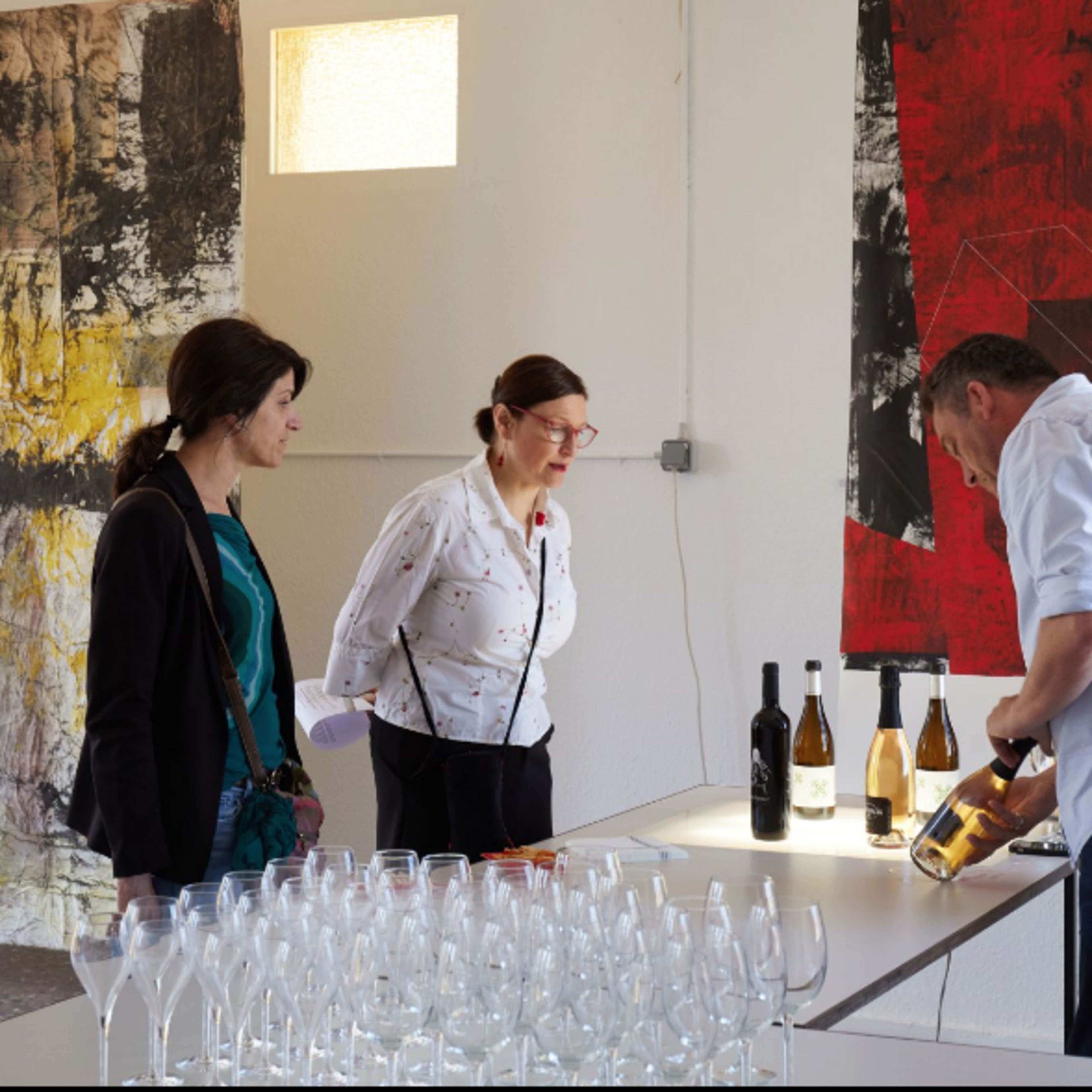 Three people observe a man as he sets up wine bottles on a table in an art-filled room while glasses are arranged in the foreground.