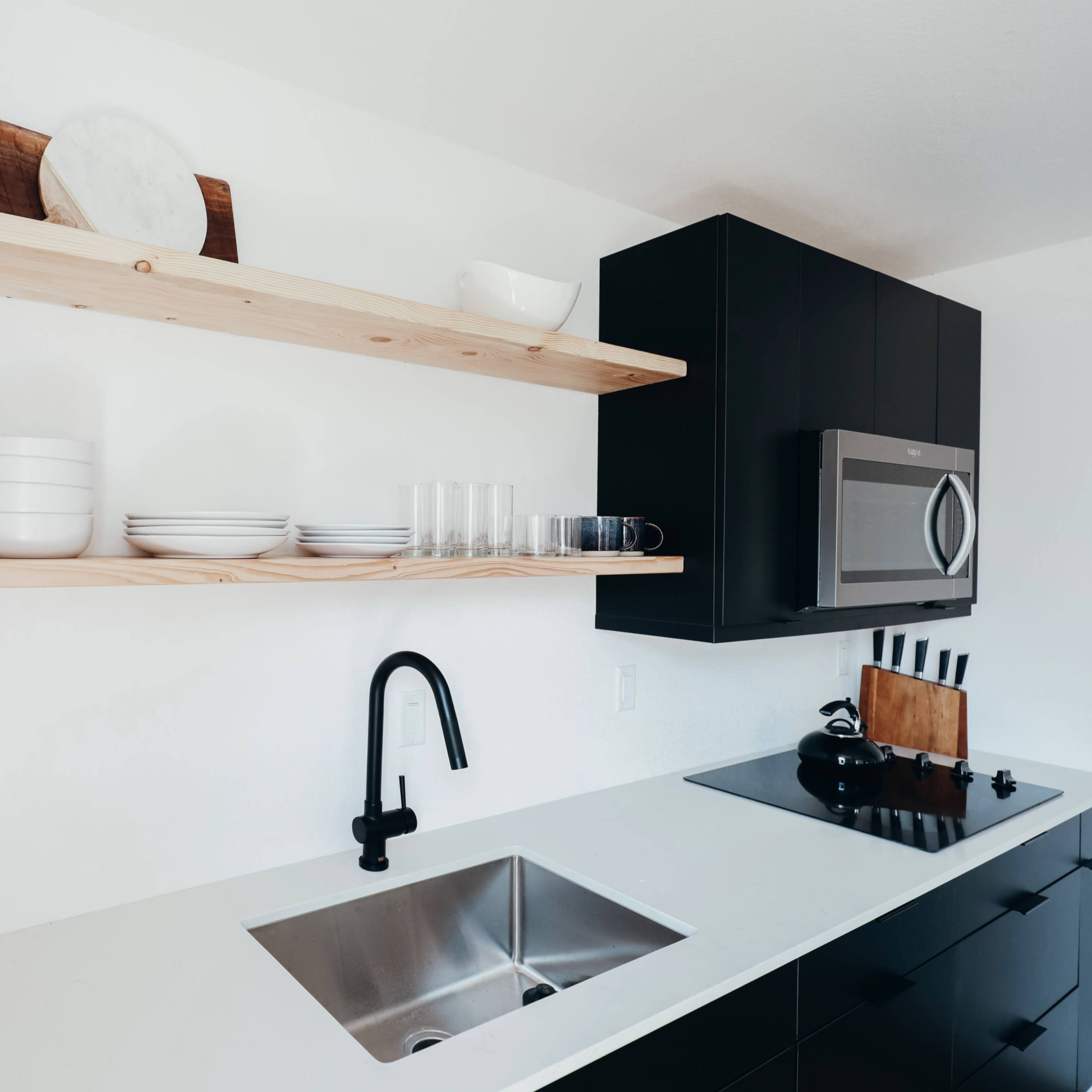 A modern kitchen features black cabinetry, open wooden shelves displaying dishes and glasses, and a stainless steel sink with a pull-down faucet.