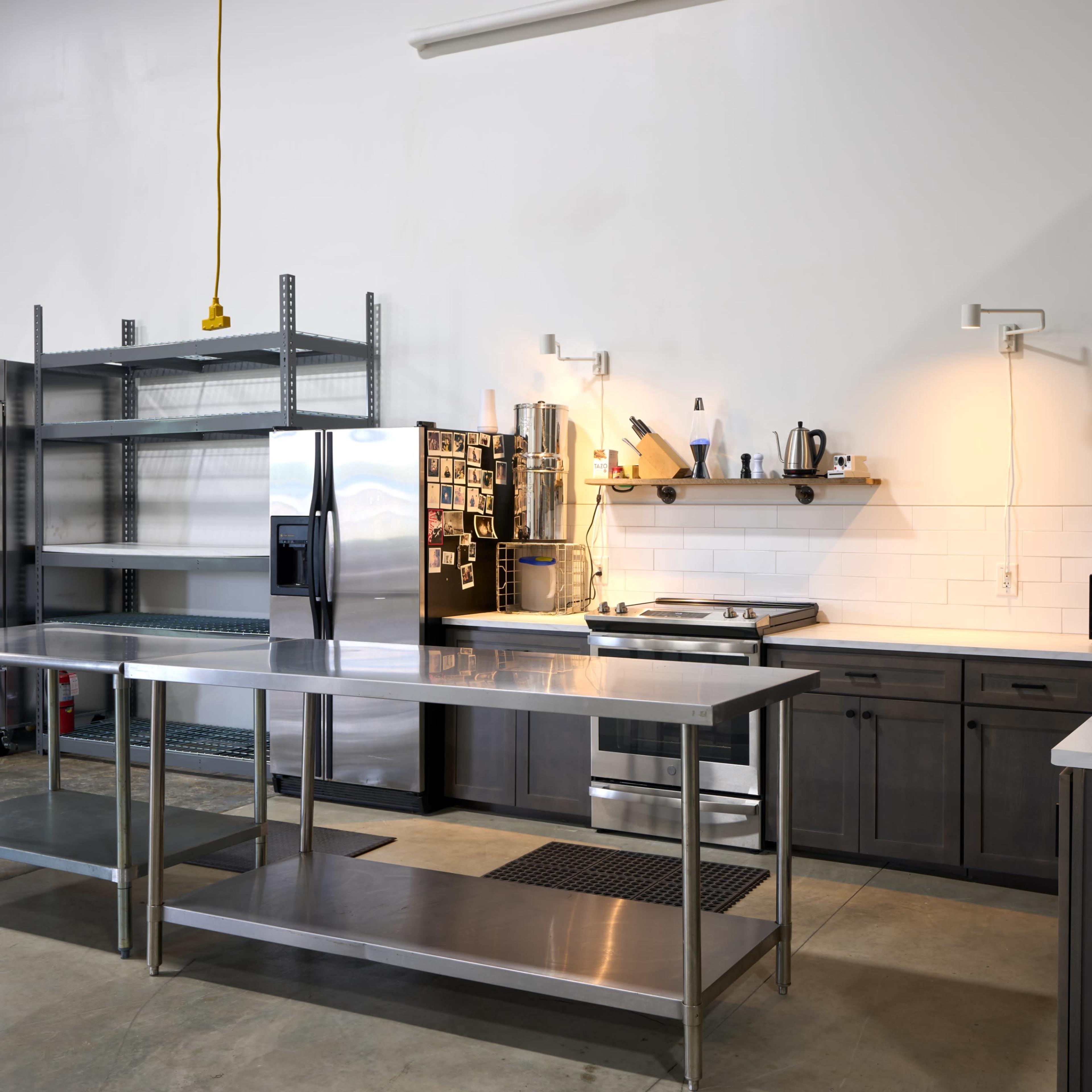 A modern kitchen with stainless steel countertops, dark cabinetry, and shelving units, featuring basic appliances and utensils.