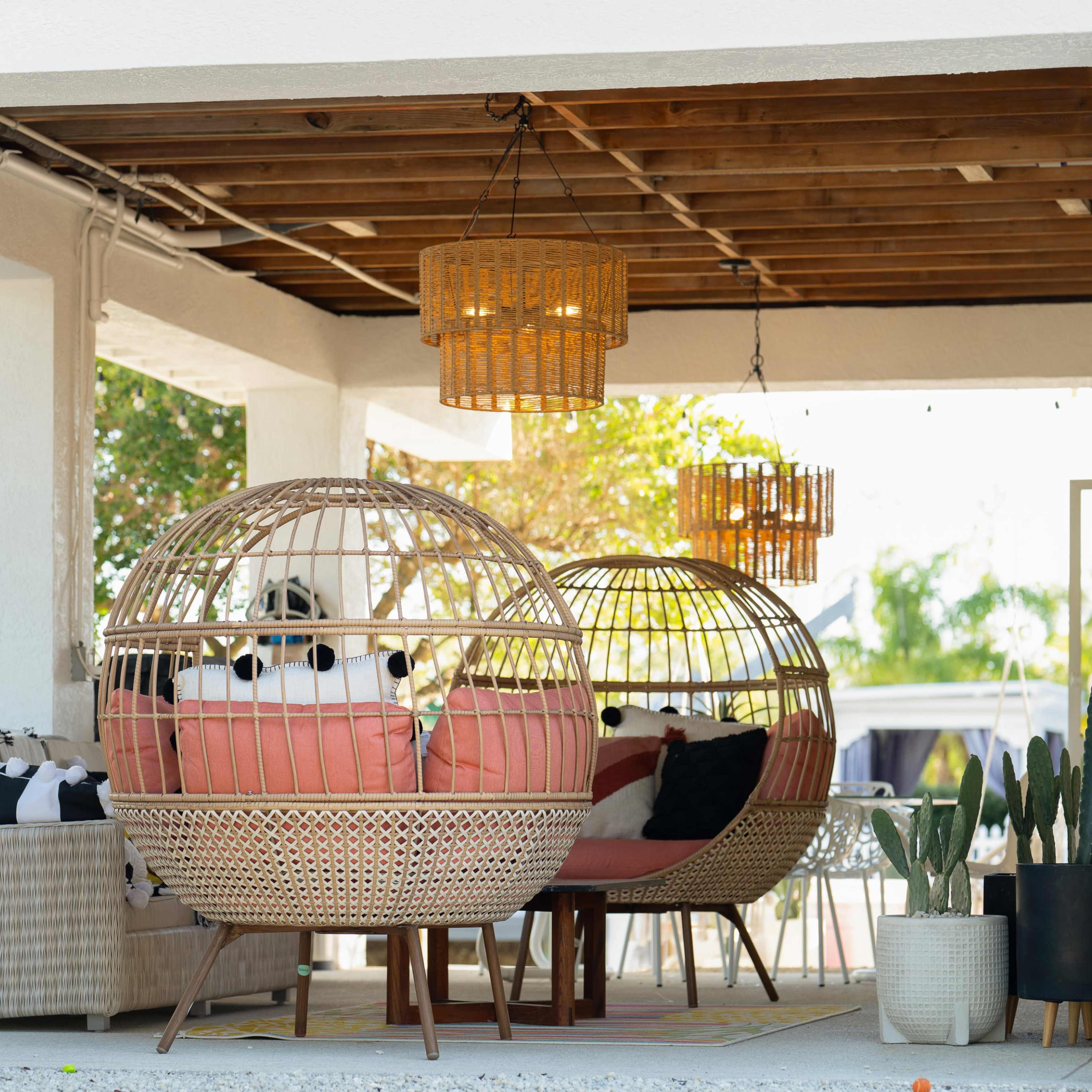 The scene features two round wicker chairs with cushions under a covered patio, surrounded by greenery and decorated with pendant lights.