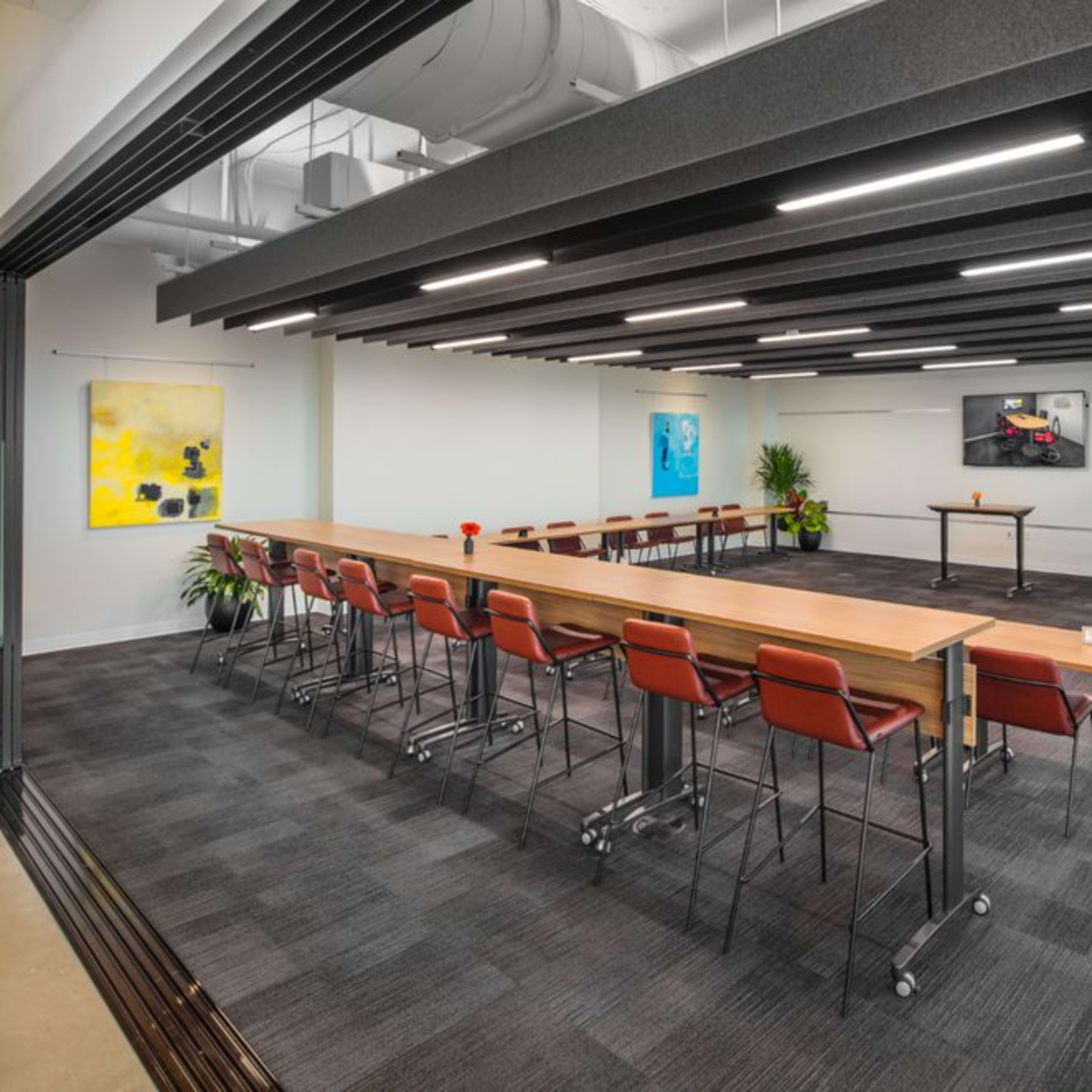 A modern conference room with a long rectangular table, red chairs, and a presentation screen at the front.