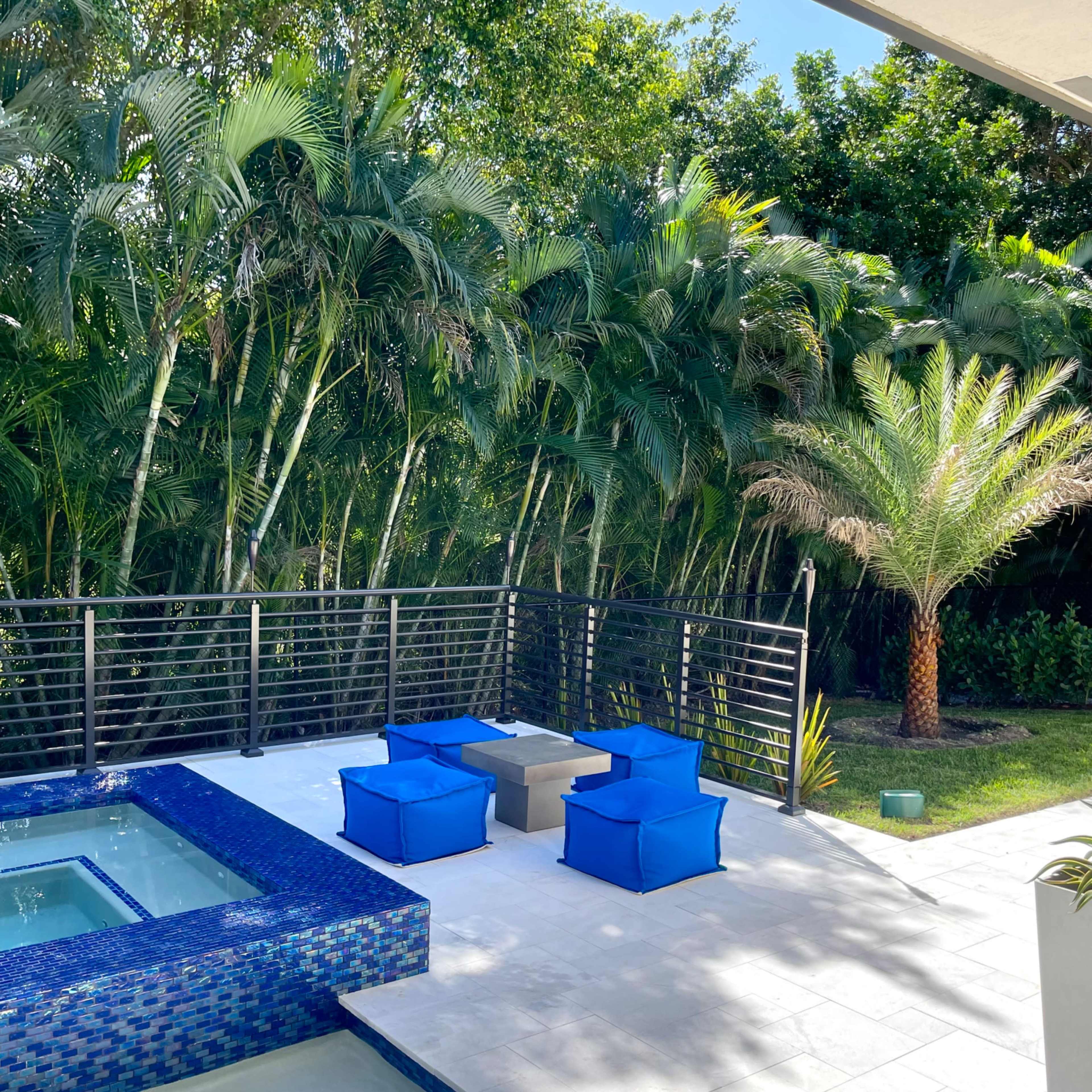 A modern outdoor space featuring a rectangular pool with a blue tiled border, accompanied by four blue lounge chairs arranged around a small table, set against a backdrop of lush greenery.