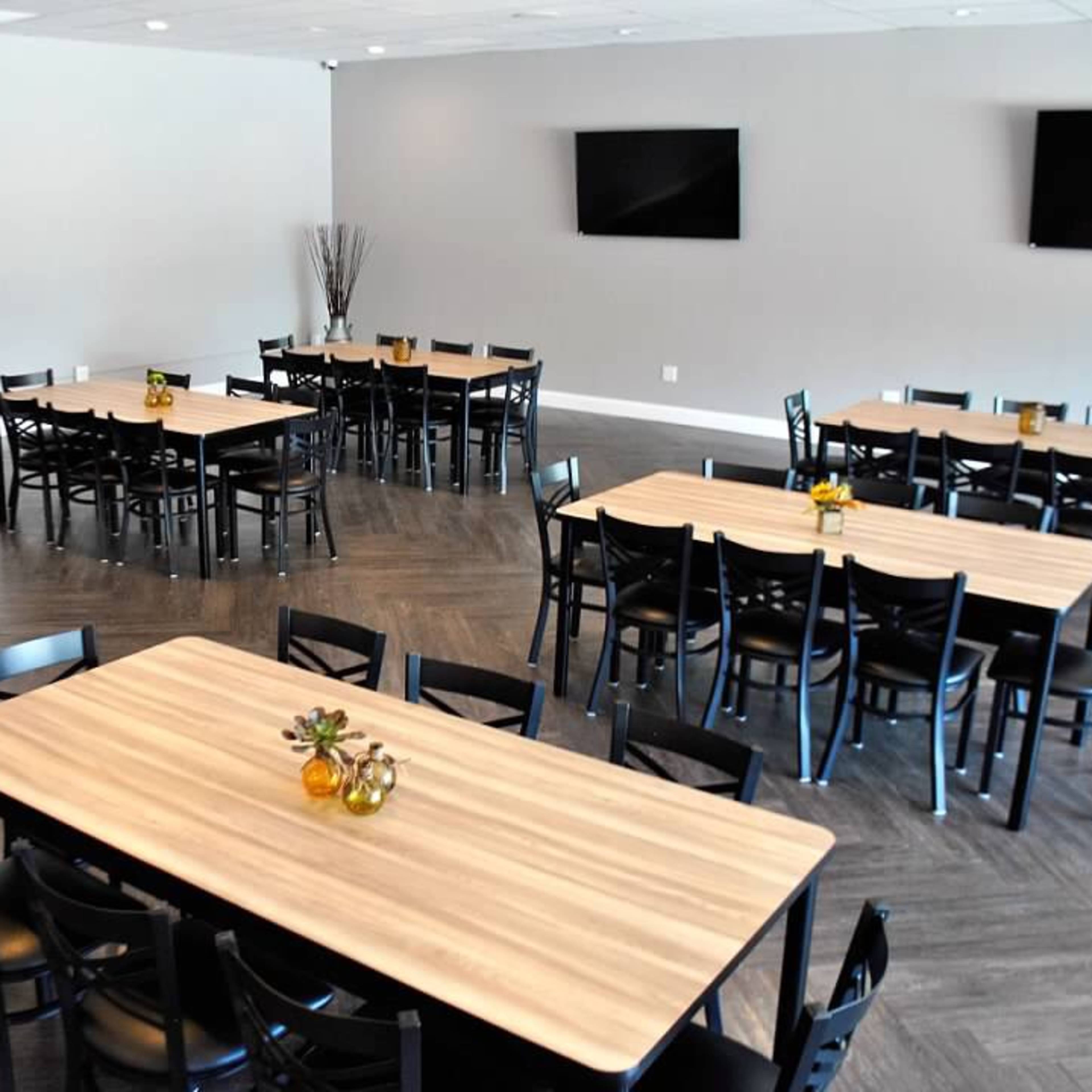 A spacious room arranged with multiple tables and chairs, featuring a modern design and two flat-screen televisions on the walls.