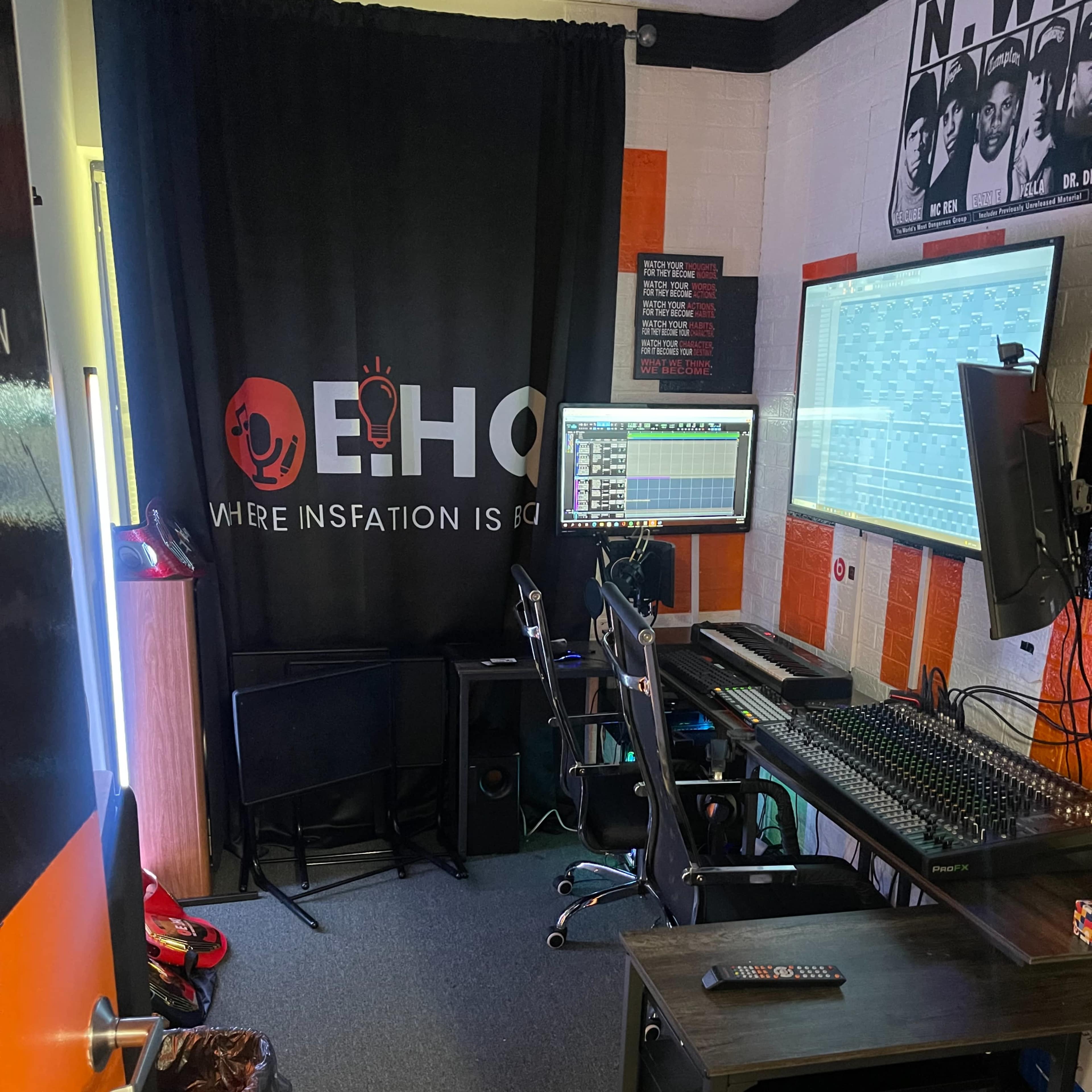 A small music studio with a mixing console, two computer monitors, and chairs arranged around a desk, all set against a backdrop of orange and black decor.