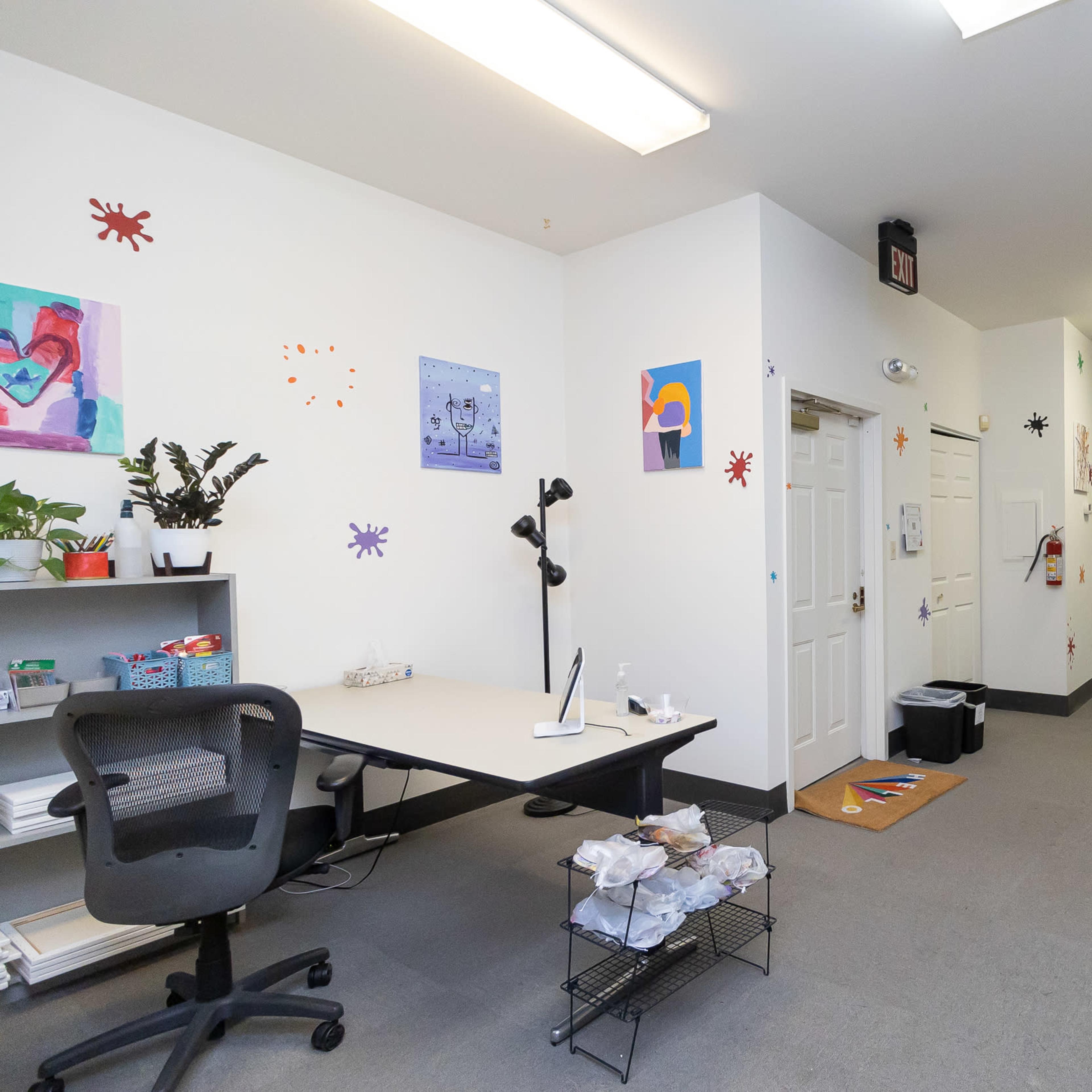 A brightly lit office space with a desk, chair, and decorative artwork on the walls, leading to a hallway with multiple doors.