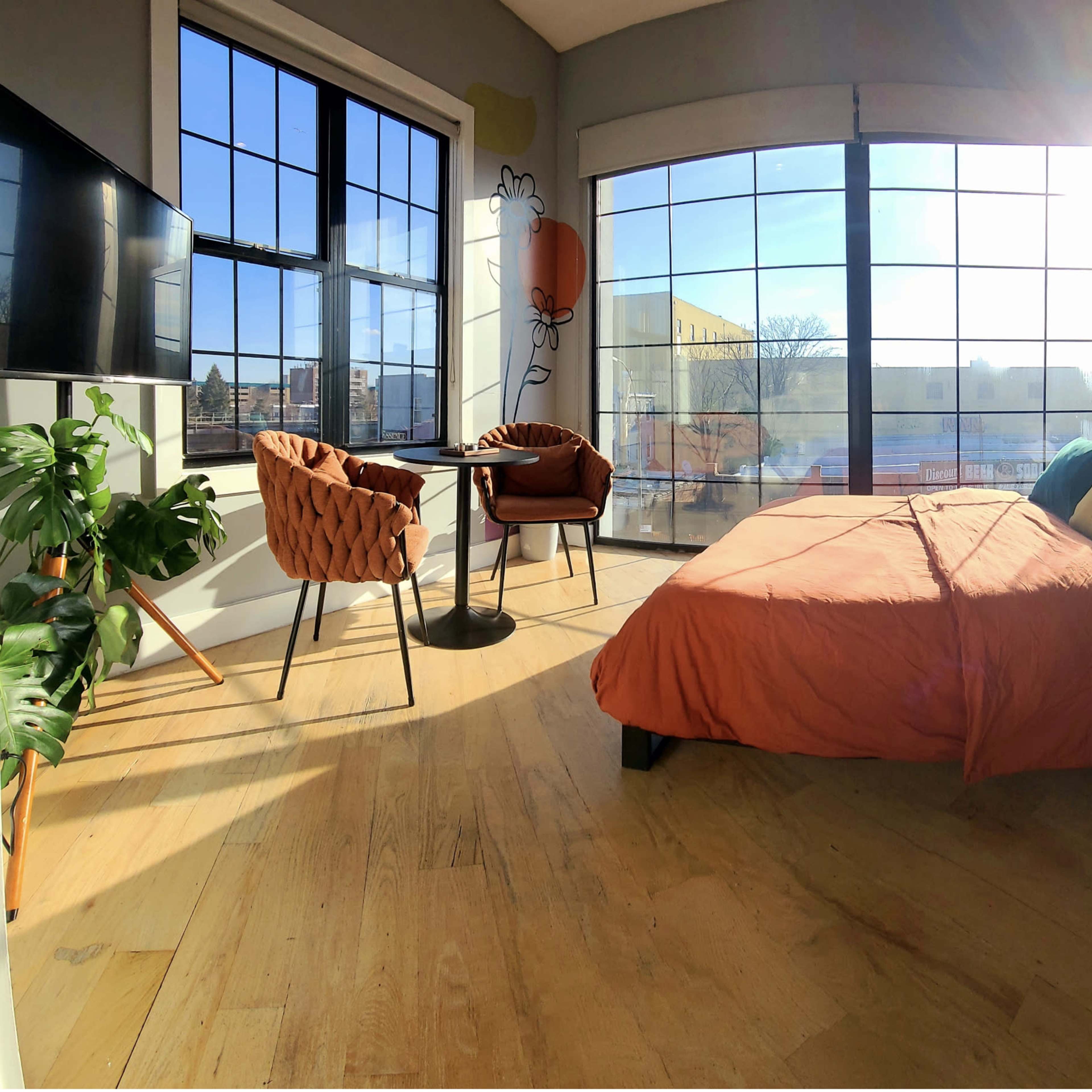 A brightly lit bedroom features a bed with an orange cover, two chairs at a small table, and large windows providing a view of the outdoors.