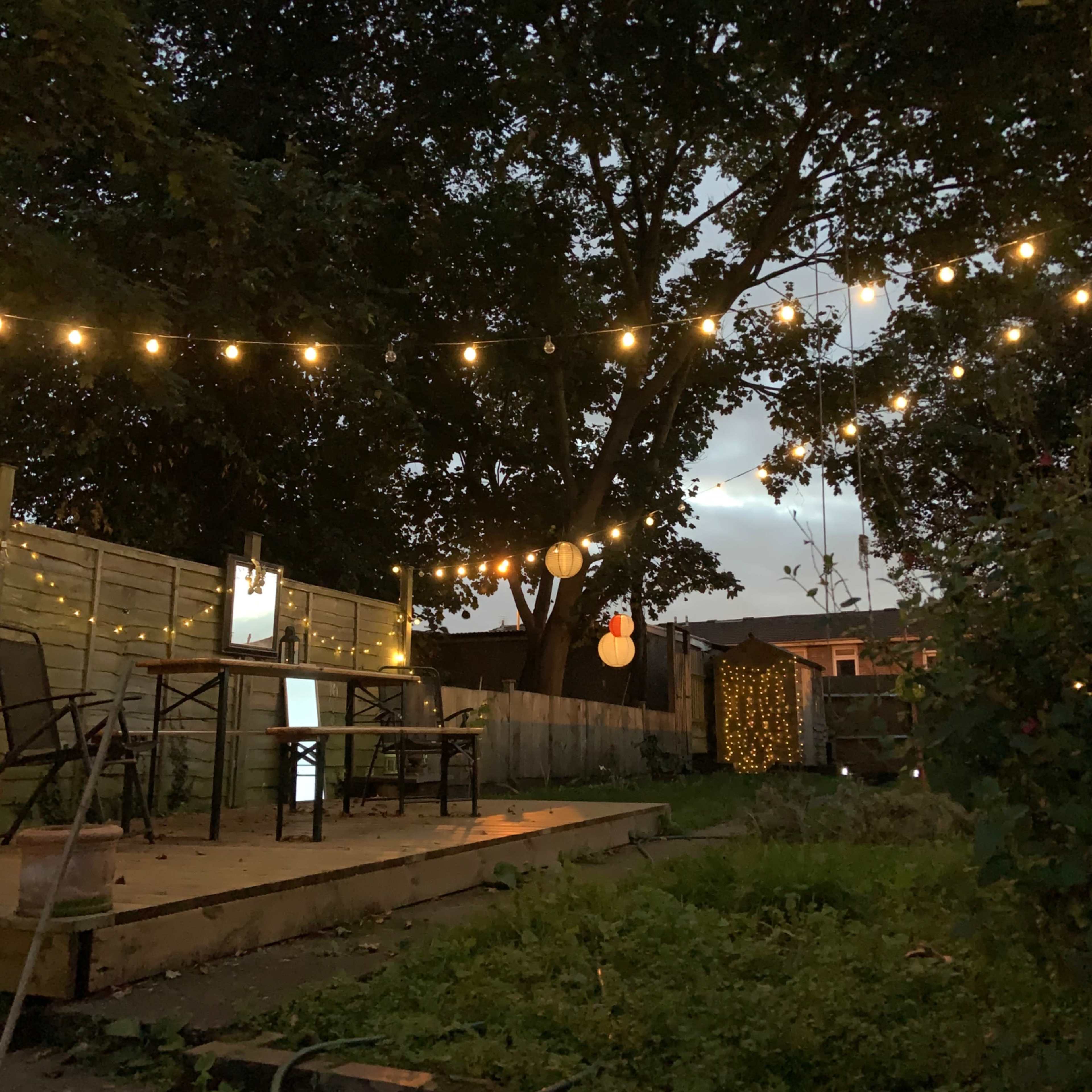 A backyard scene features a wooden deck with string lights overhead, surrounded by greenery and decorative lanterns hanging from a tree.