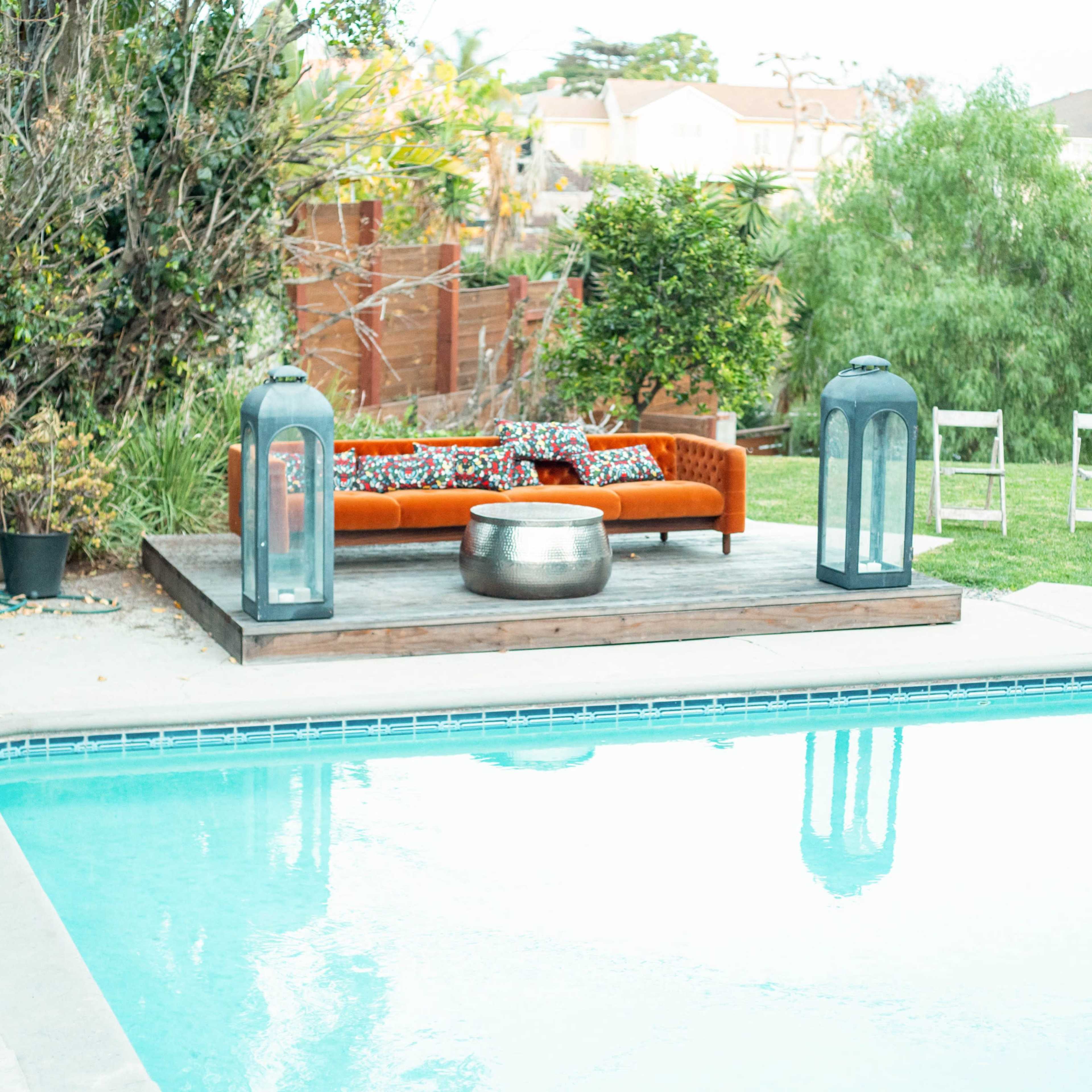 A swimming pool is surrounded by a wooden deck featuring a couch with decorative pillows, two lanterns, and a metal table, with chairs arranged nearby on the grass.