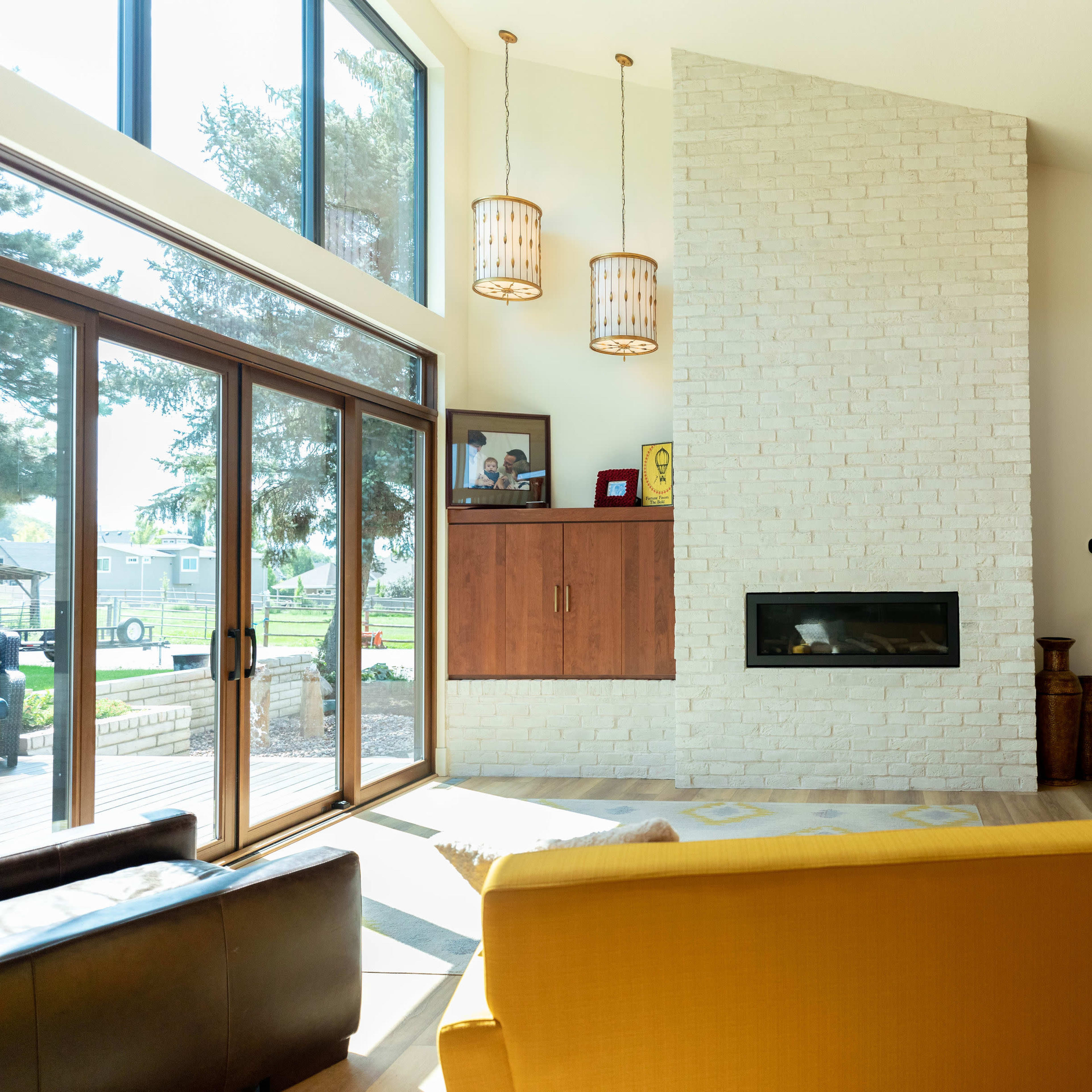 A modern living room features a yellow sofa facing a wall-mounted fireplace, large windows, and wooden ceiling beams.