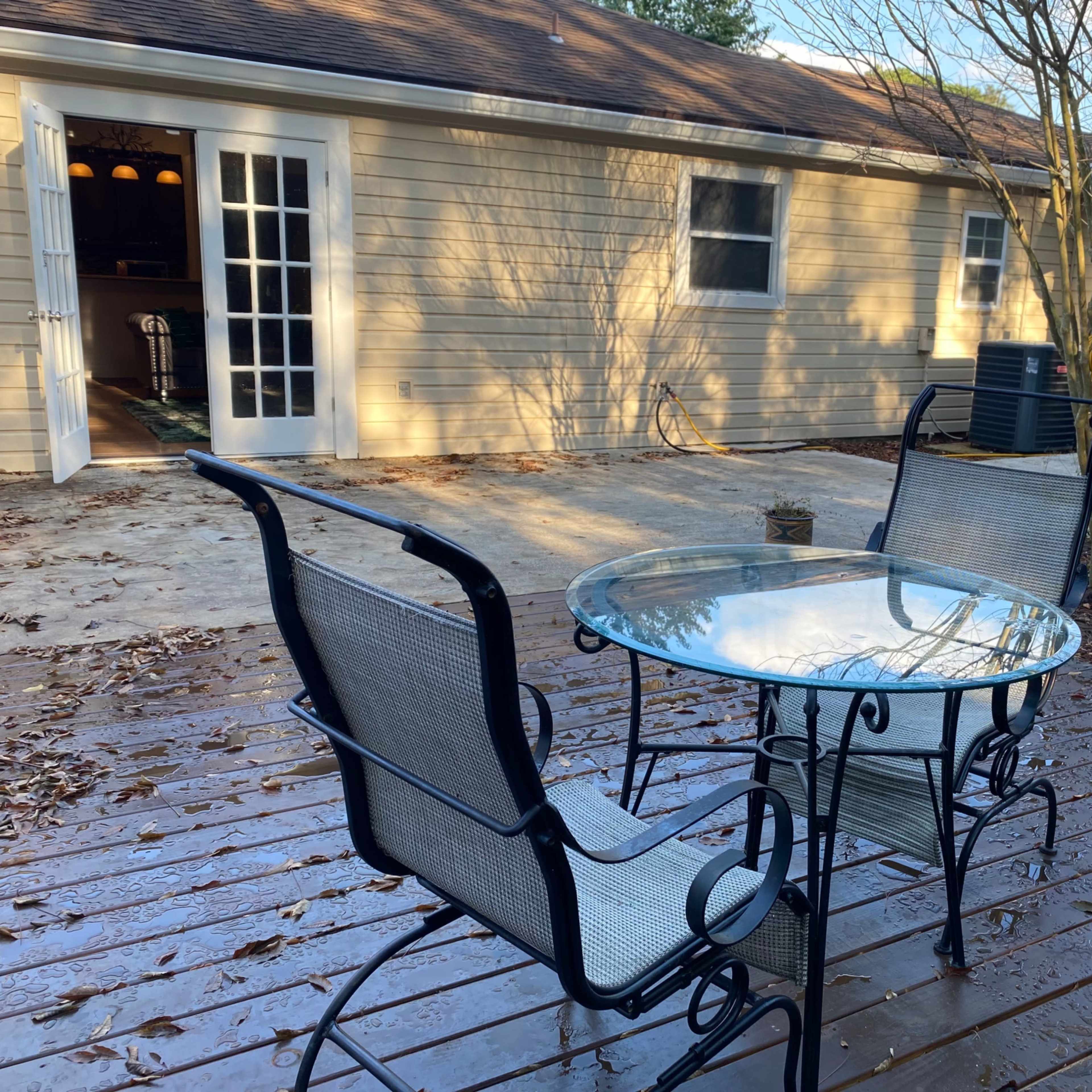 A round glass table with two metal chairs is positioned on a wooden patio, facing an exterior wall of a house.