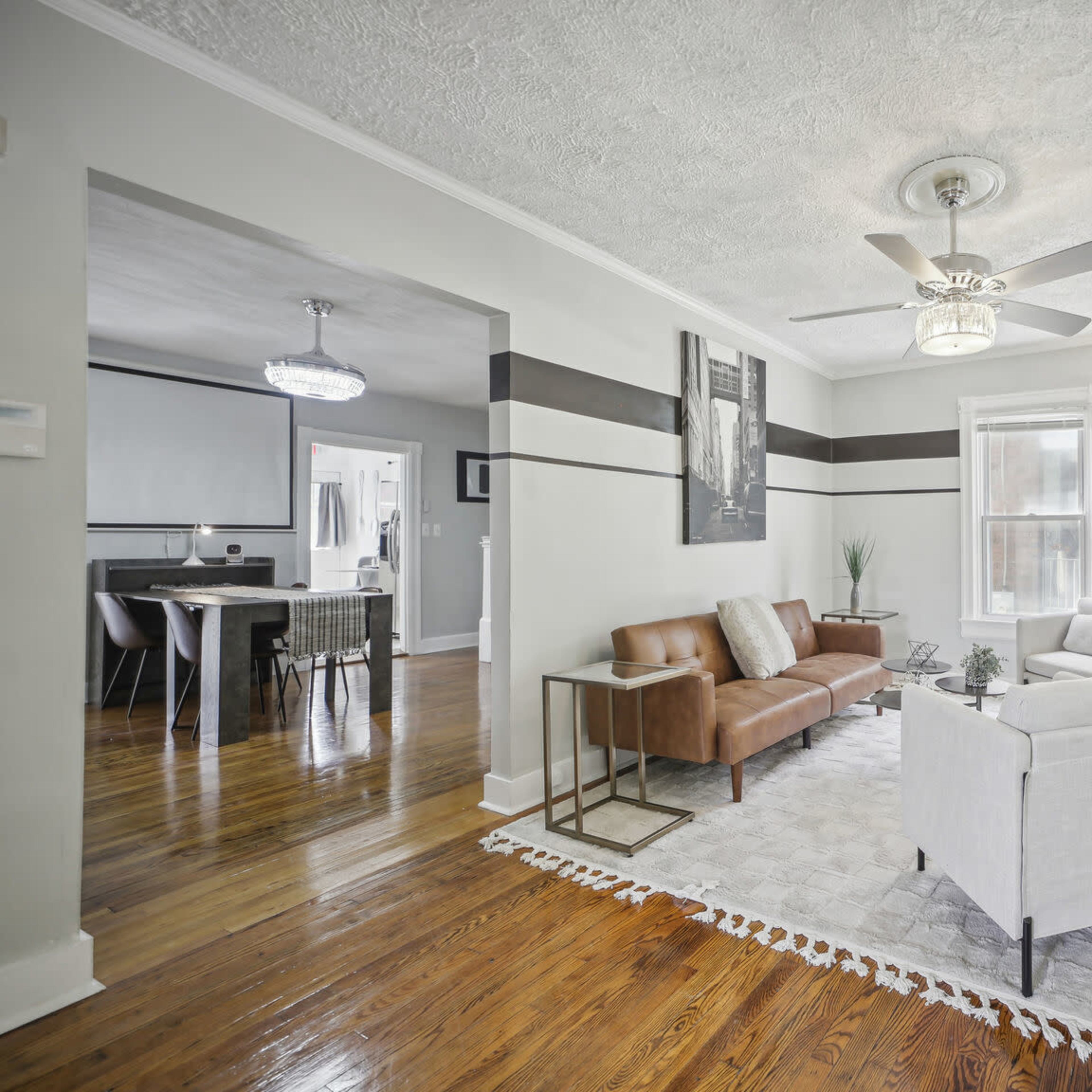 A living room with hardwood floors, a brown leather sofa, white armchairs, and a ceiling fan, leading into a dining area visible beyond.
