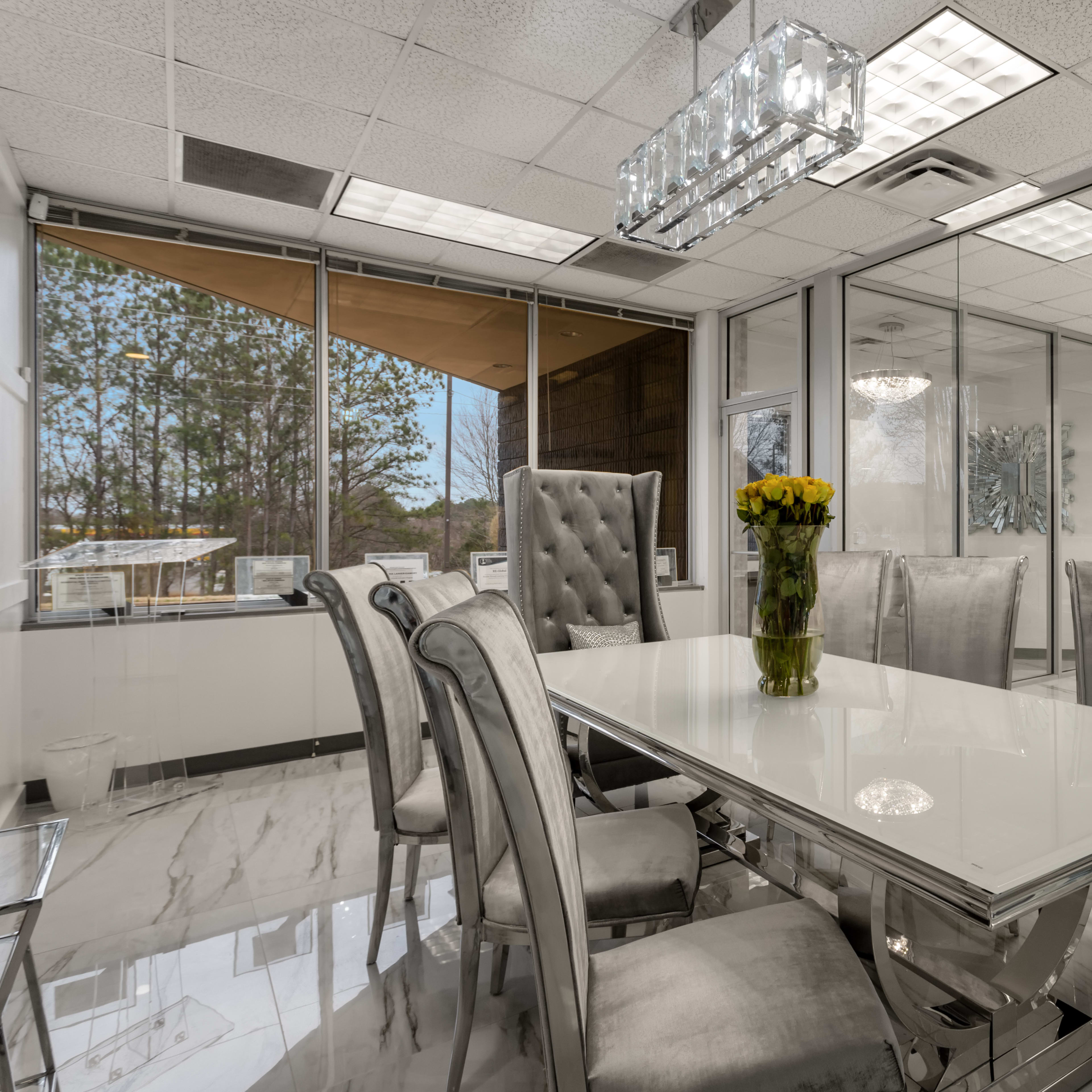 A modern glass-walled dining area features a long white table surrounded by upholstered chairs and a floral centerpiece.