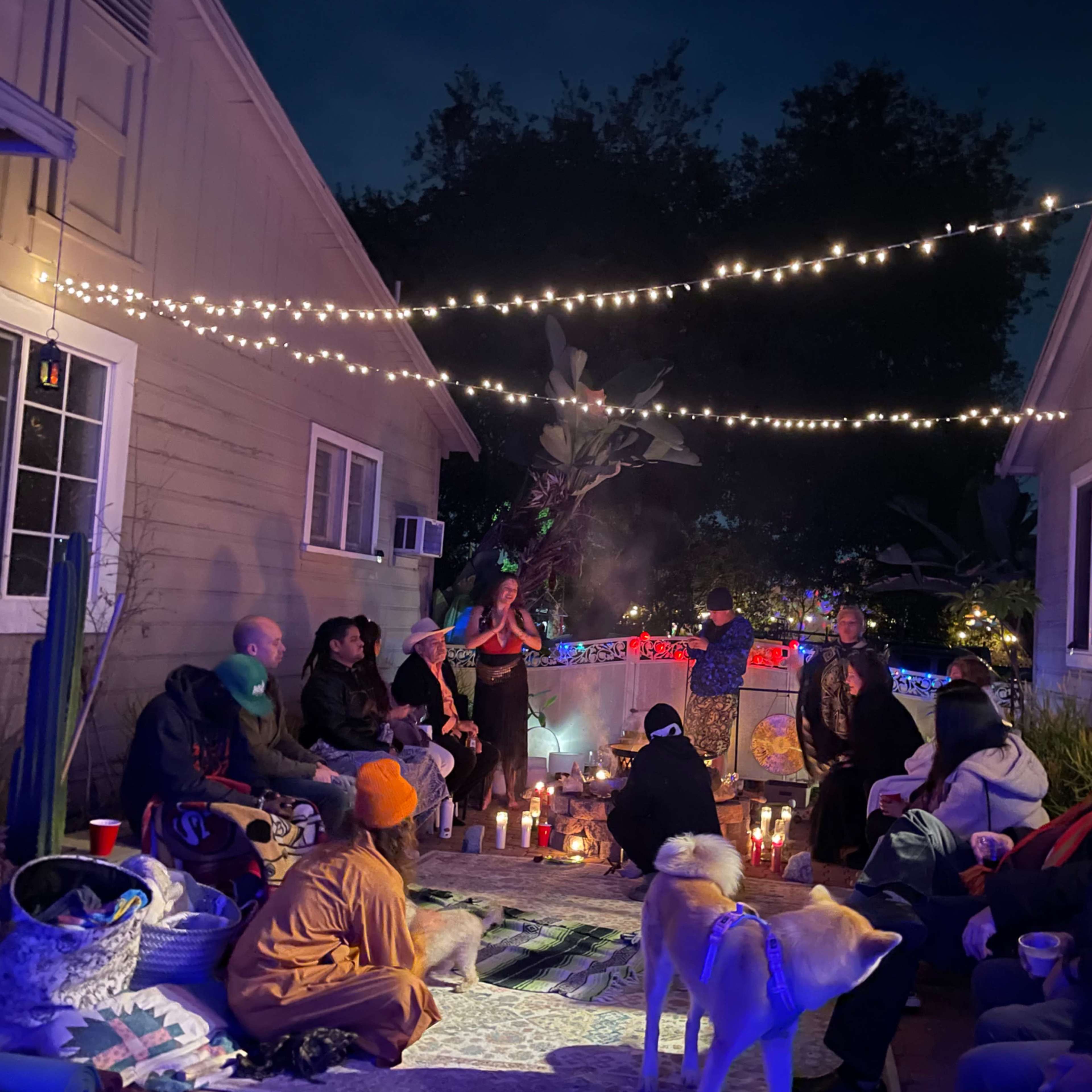 A group of people sits on a patio surrounded by lights and candles, with a performer in the center, during a nighttime gathering.
