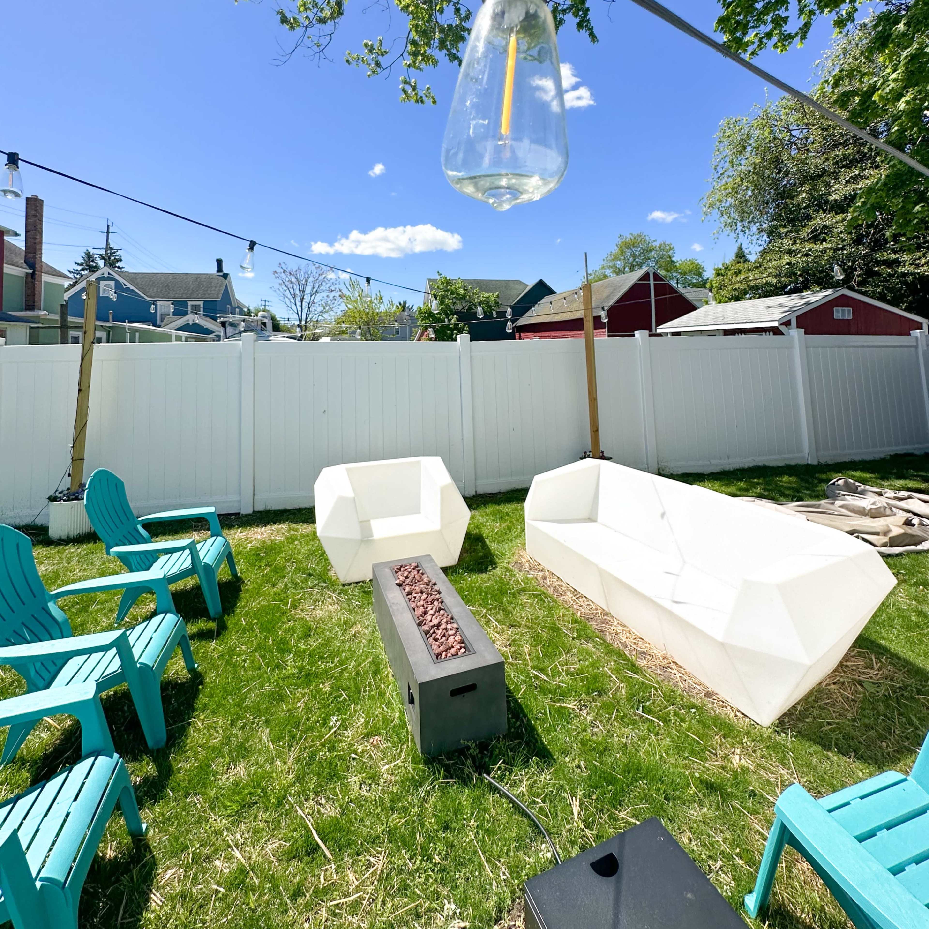A backyard seating area features modern outdoor furniture arranged around a fire pit, with a light bulb hanging overhead and a white fence in the background.
