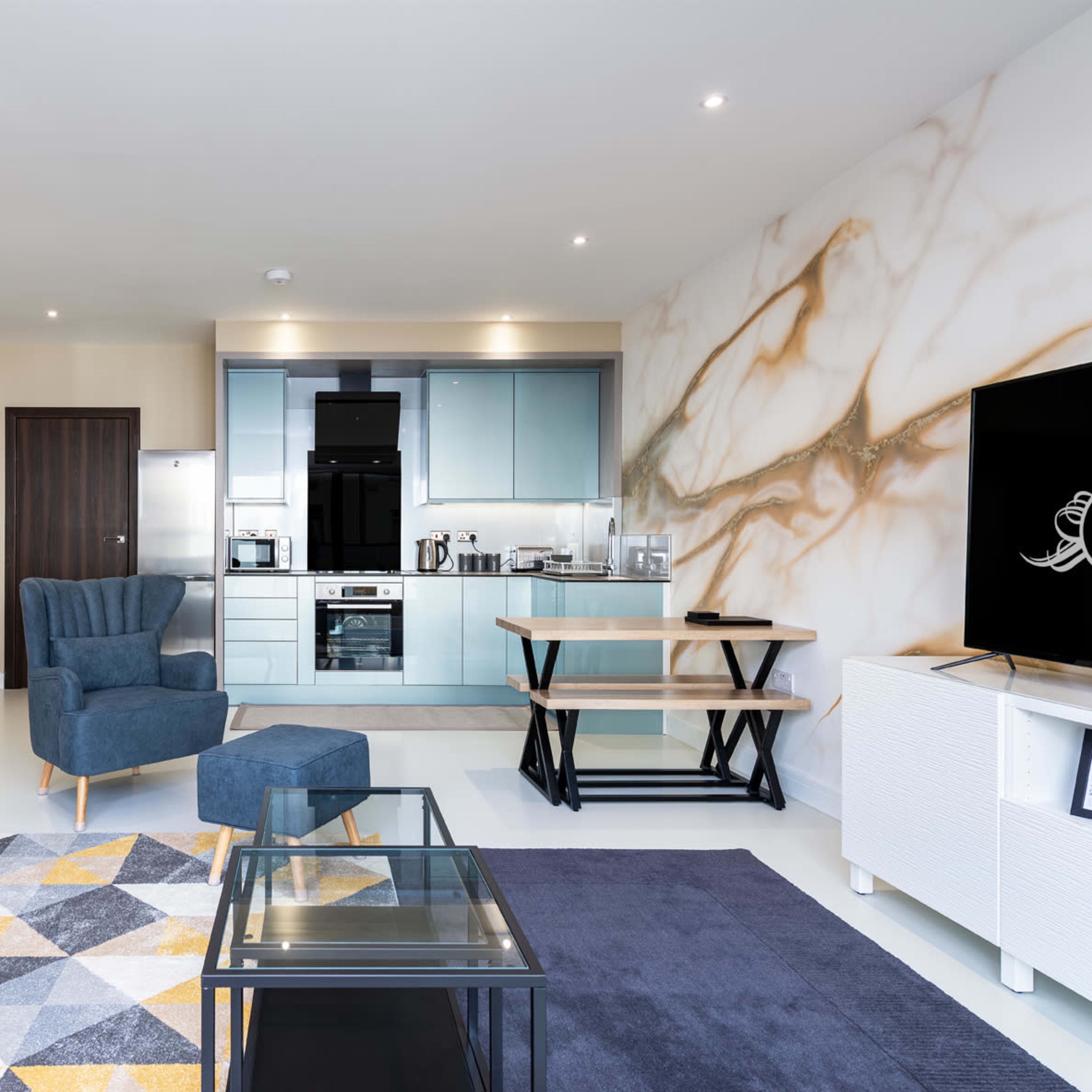 A modern living space featuring a blue sofa, an armchair, a dining table, and a kitchen with light blue cabinetry.