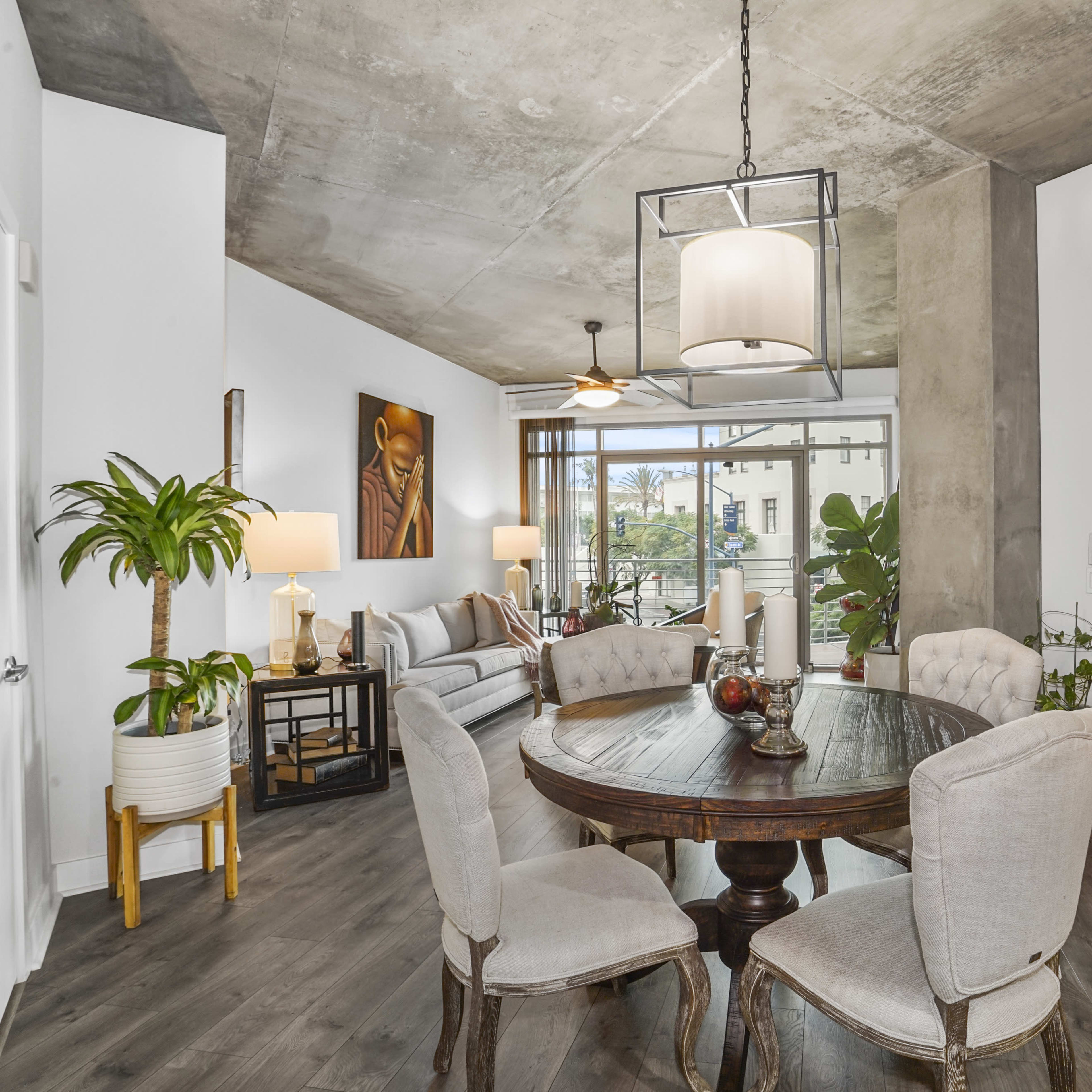 A modern living space features a round dining table surrounded by upholstered chairs, with a seating area visible in the background and large windows letting in natural light.